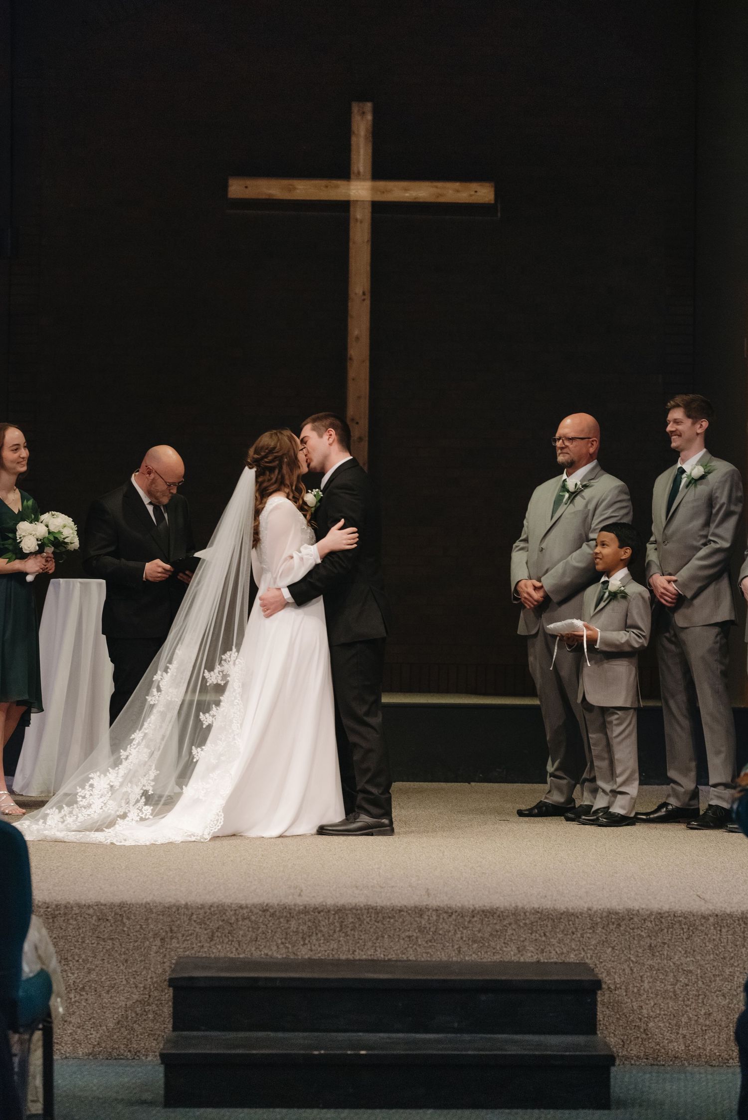 Bride and groom share first kiss at church altar with wooden cross, wedding party standing alongside.