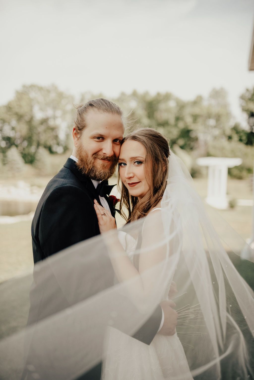 Bride and groom posing together outdoors on wedding day, bride wearing veil flowing in wind.