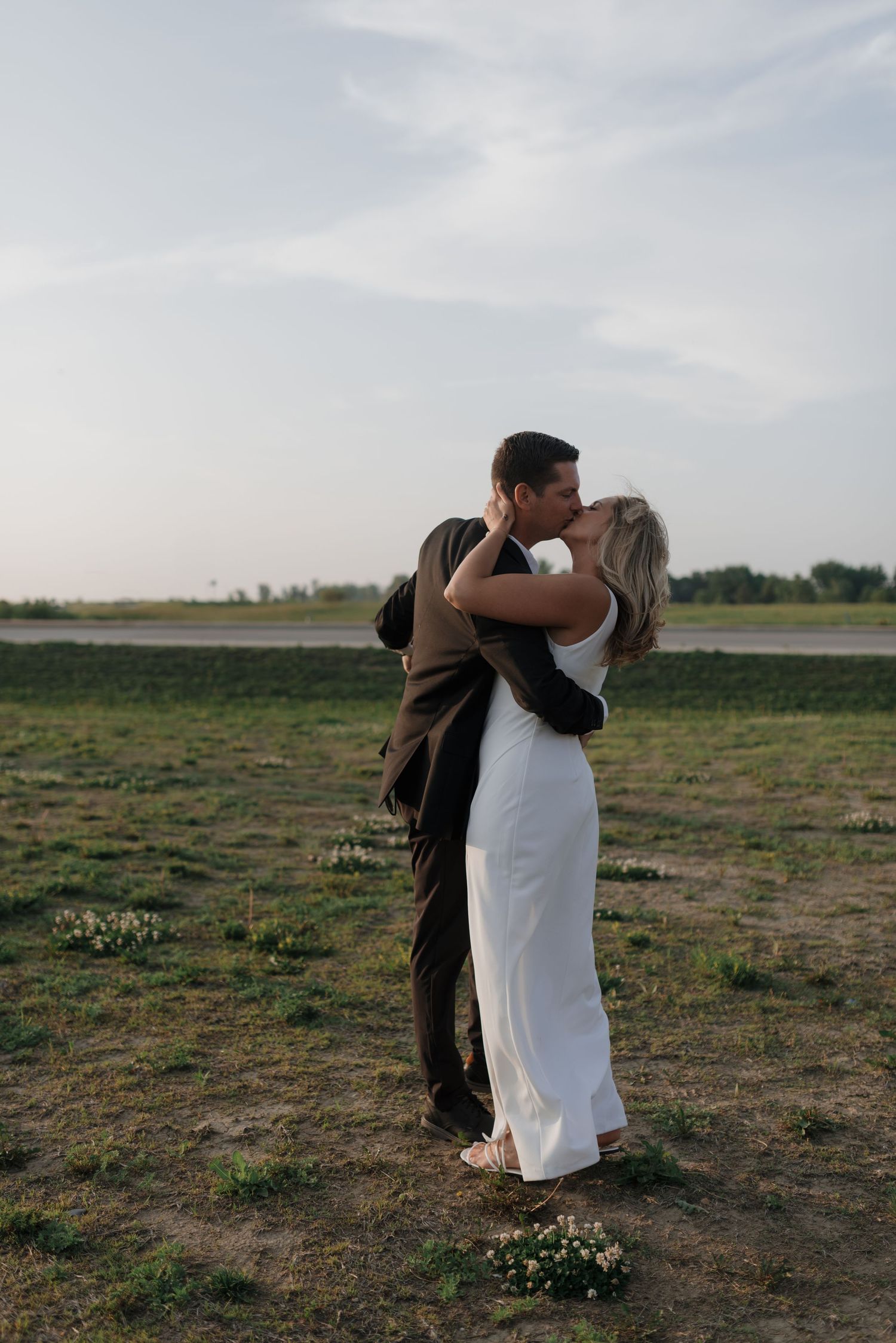 Newlyweds share passionate kiss in open green field during golden hour wedding portraits.