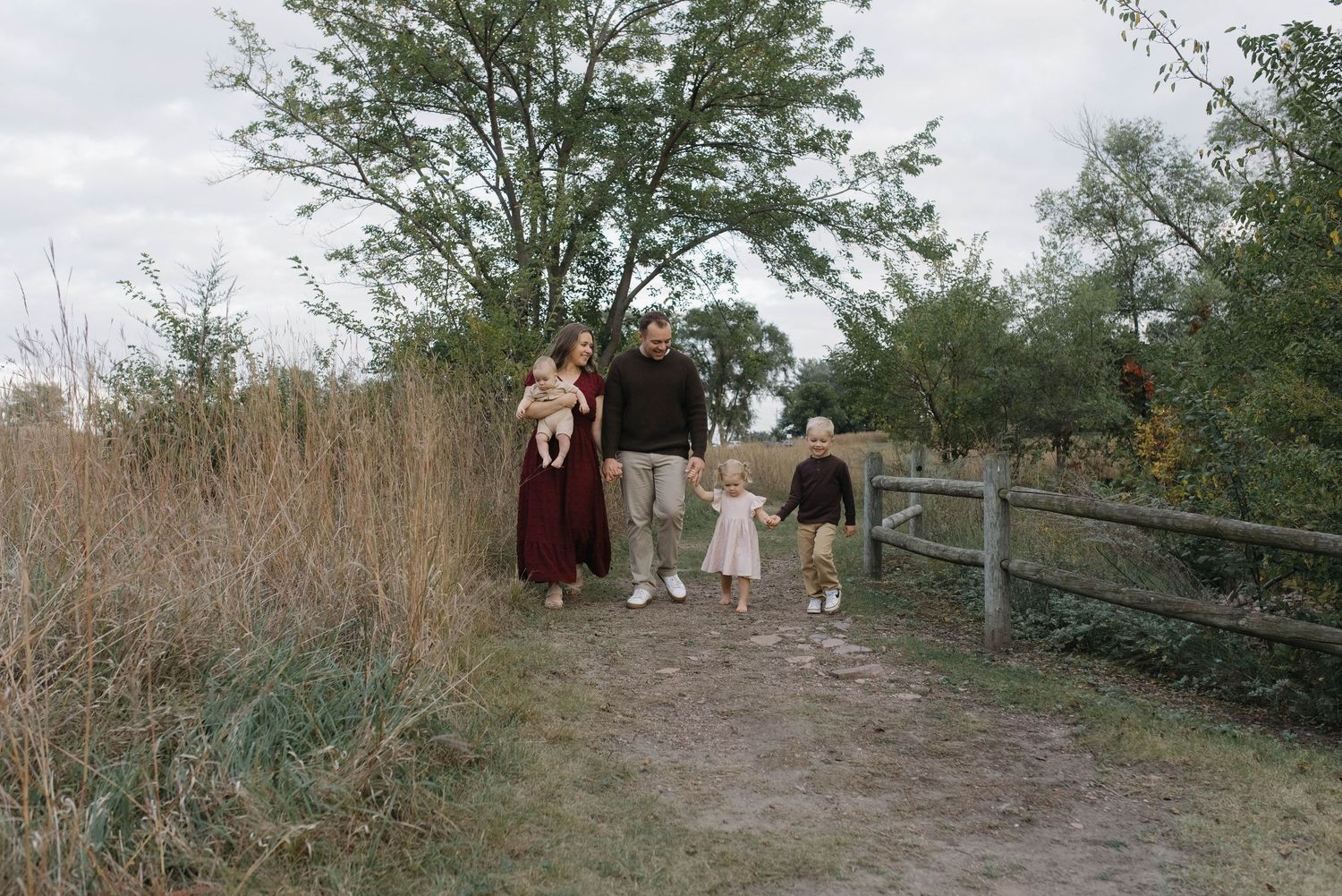 Young family strolling along a scenic dirt path bordered by tall grass and a wooden fence.