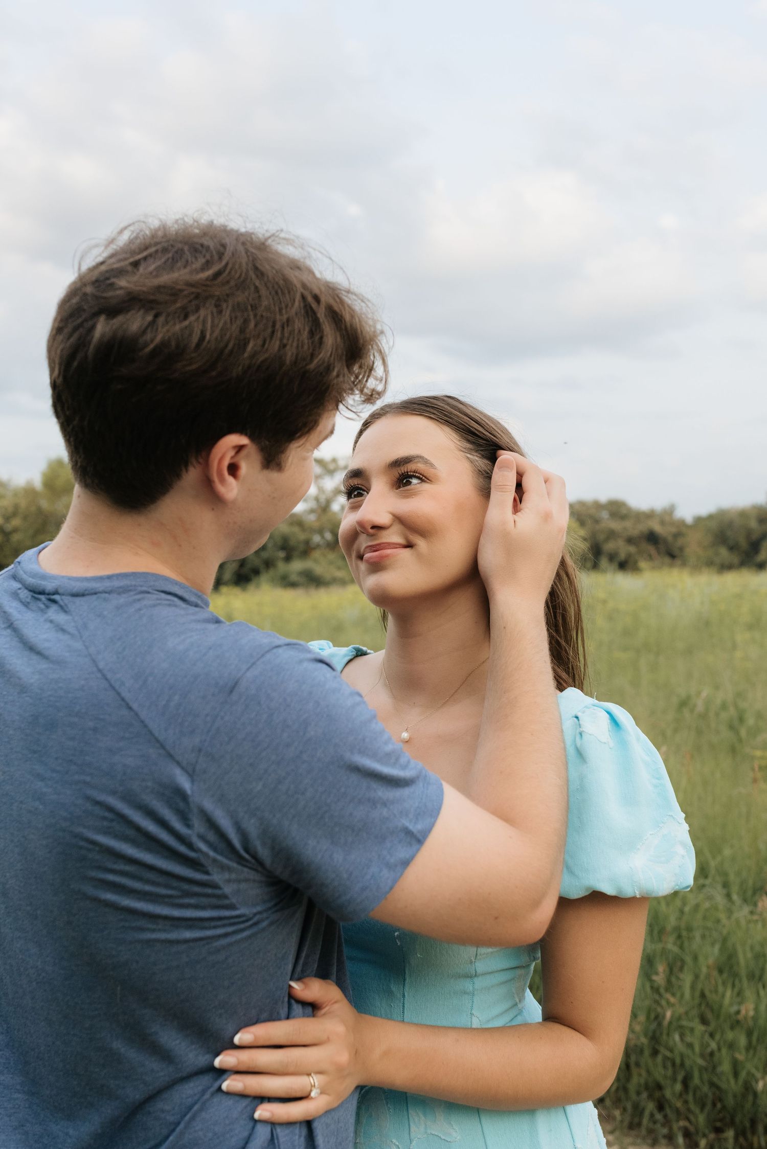 A couple shares a tender moment outdoors, man gently brushing woman's hair as she smiles in a teal dress.