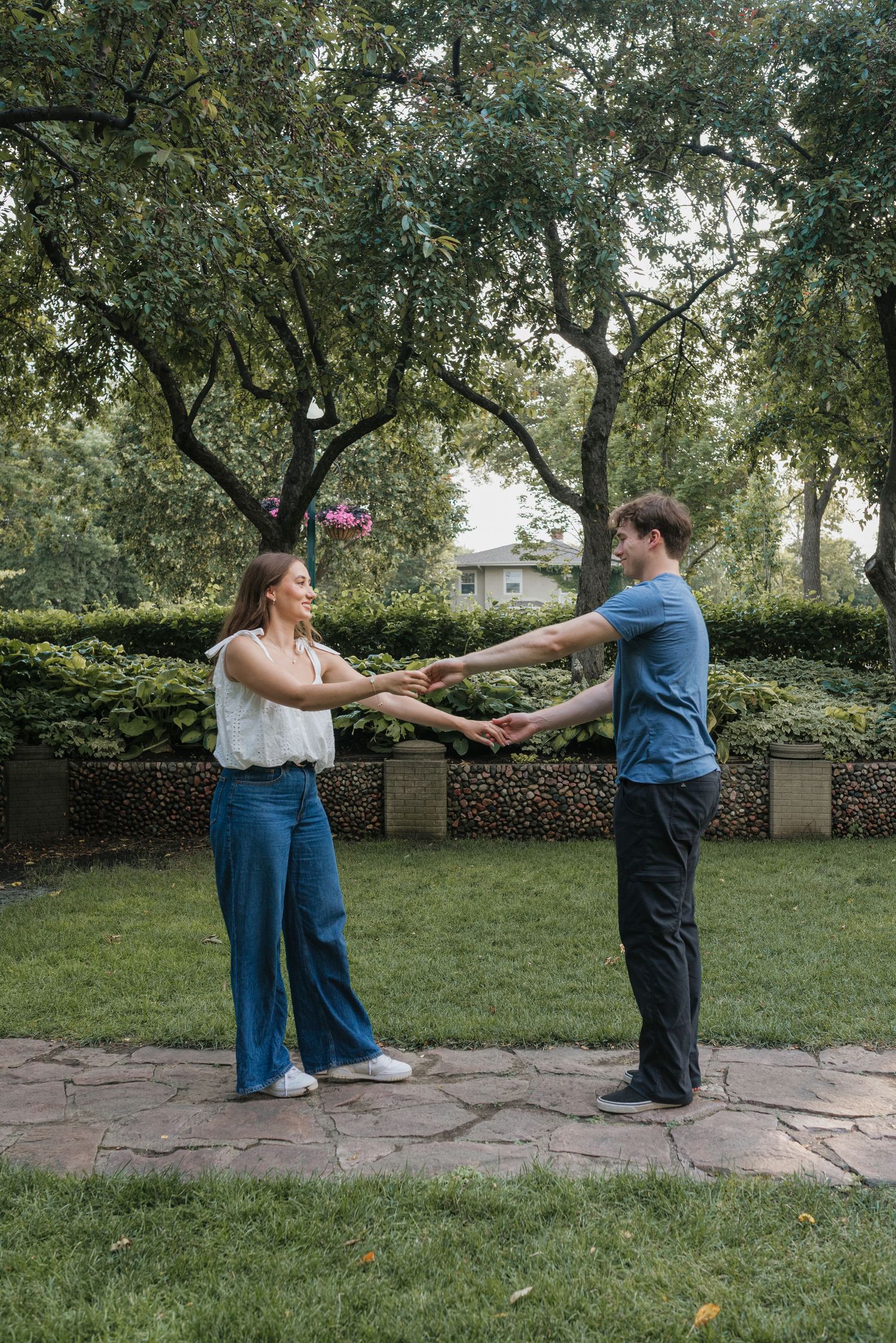 A couple holds hands and dances together on a stone path in a lush green park setting.