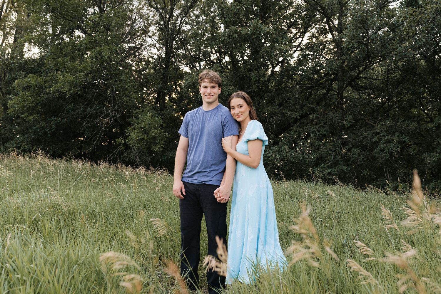 A young couple poses together in a lush green meadow, the woman wearing a light blue dress.