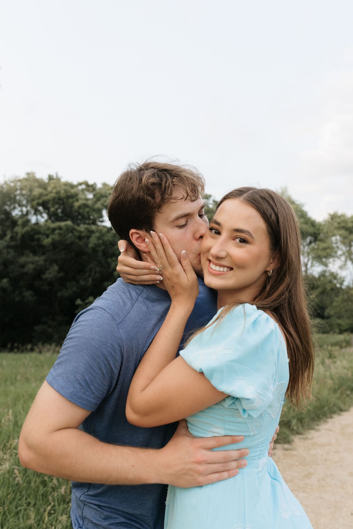 A young couple sharing a romantic moment outdoors, man kissing woman's cheek while she smiles in a light blue dress.