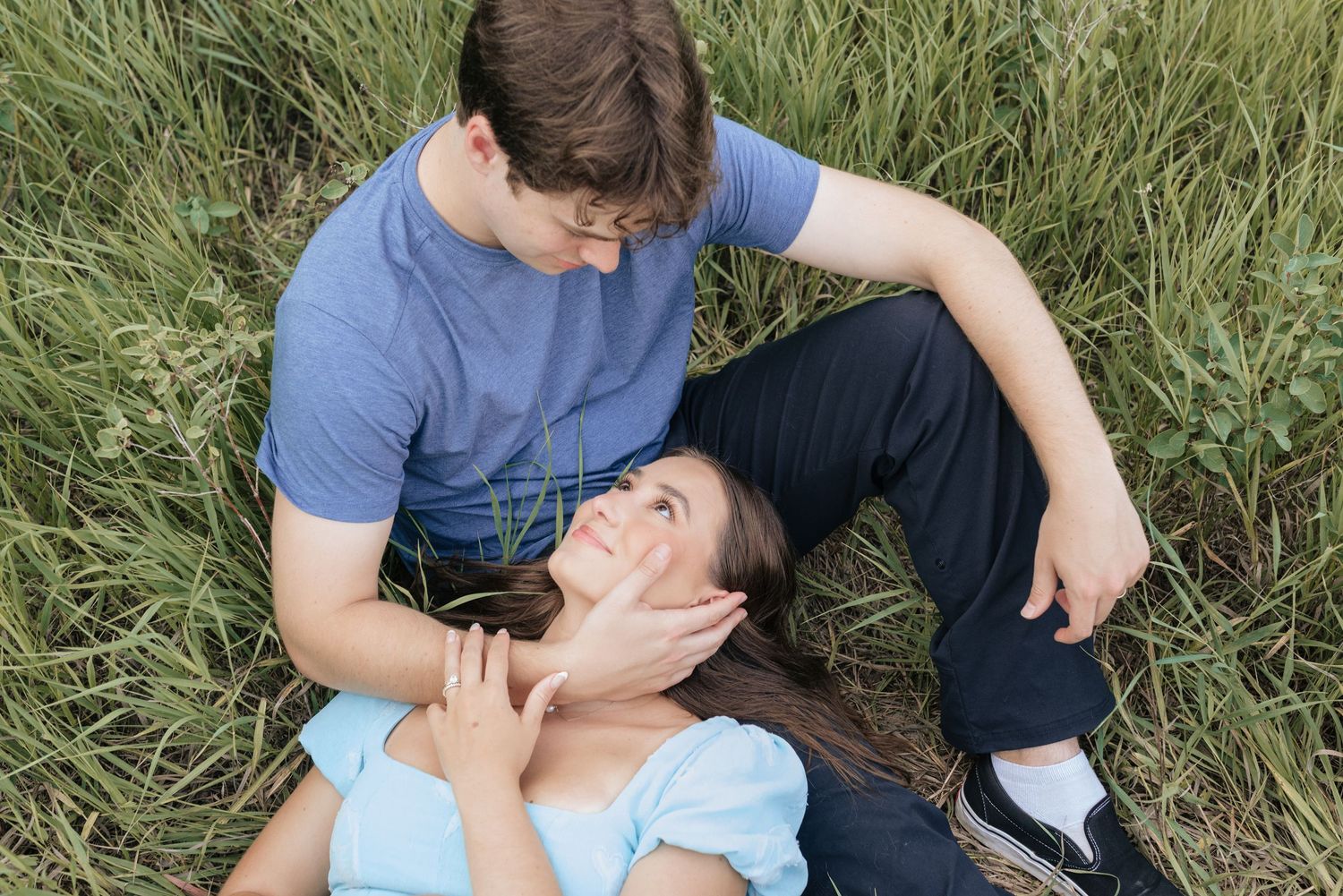 Young couple sitting in tall green grass, woman lying in man's lap gazing up at him romantically.