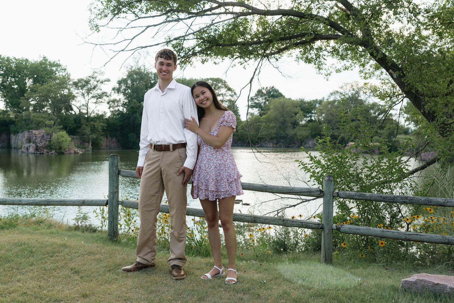 A young couple poses together near a wooden fence by a serene river, surrounded by lush green trees.
