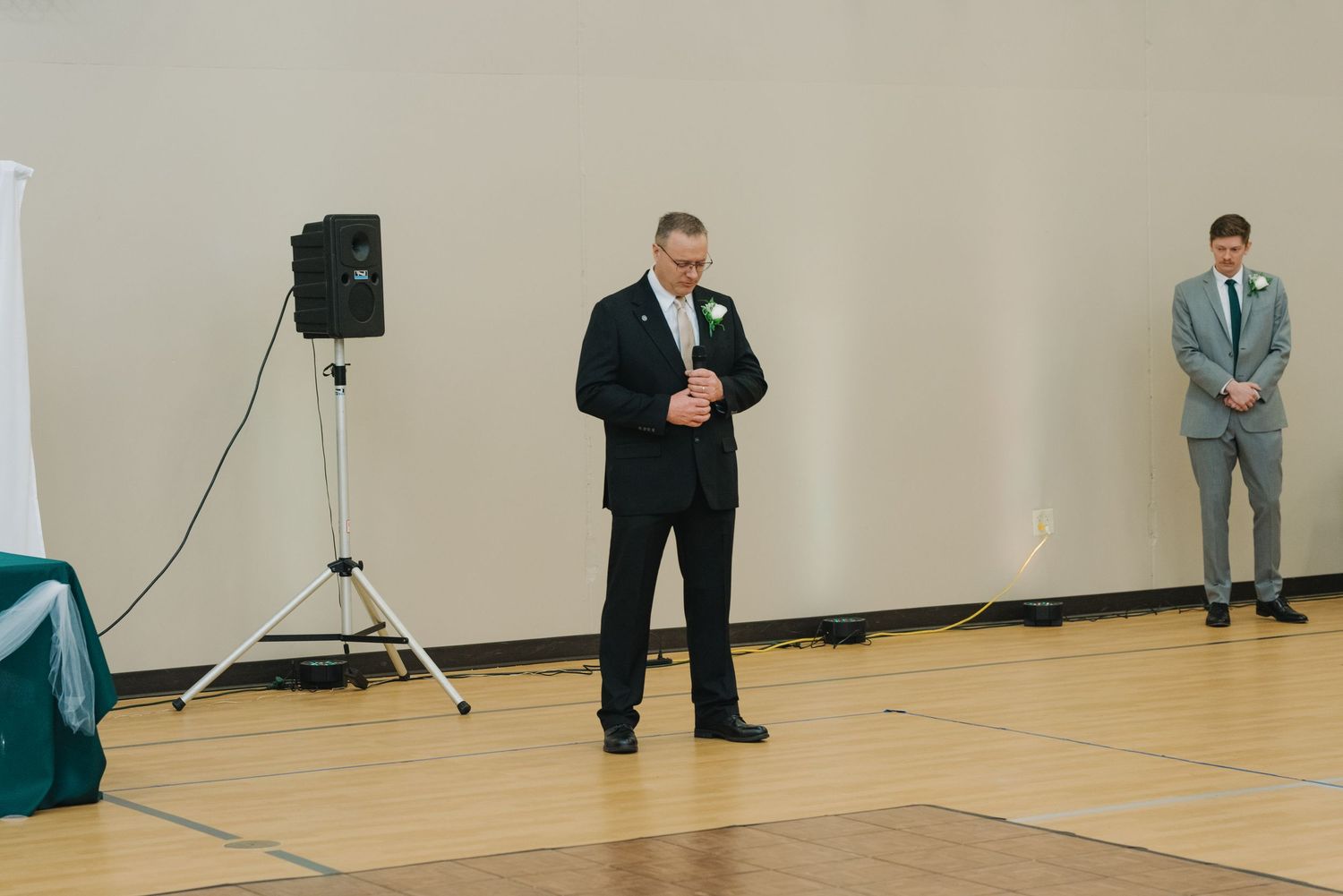 Man in black suit with boutonniere giving a speech at a wedding reception in a gymnasium venue.