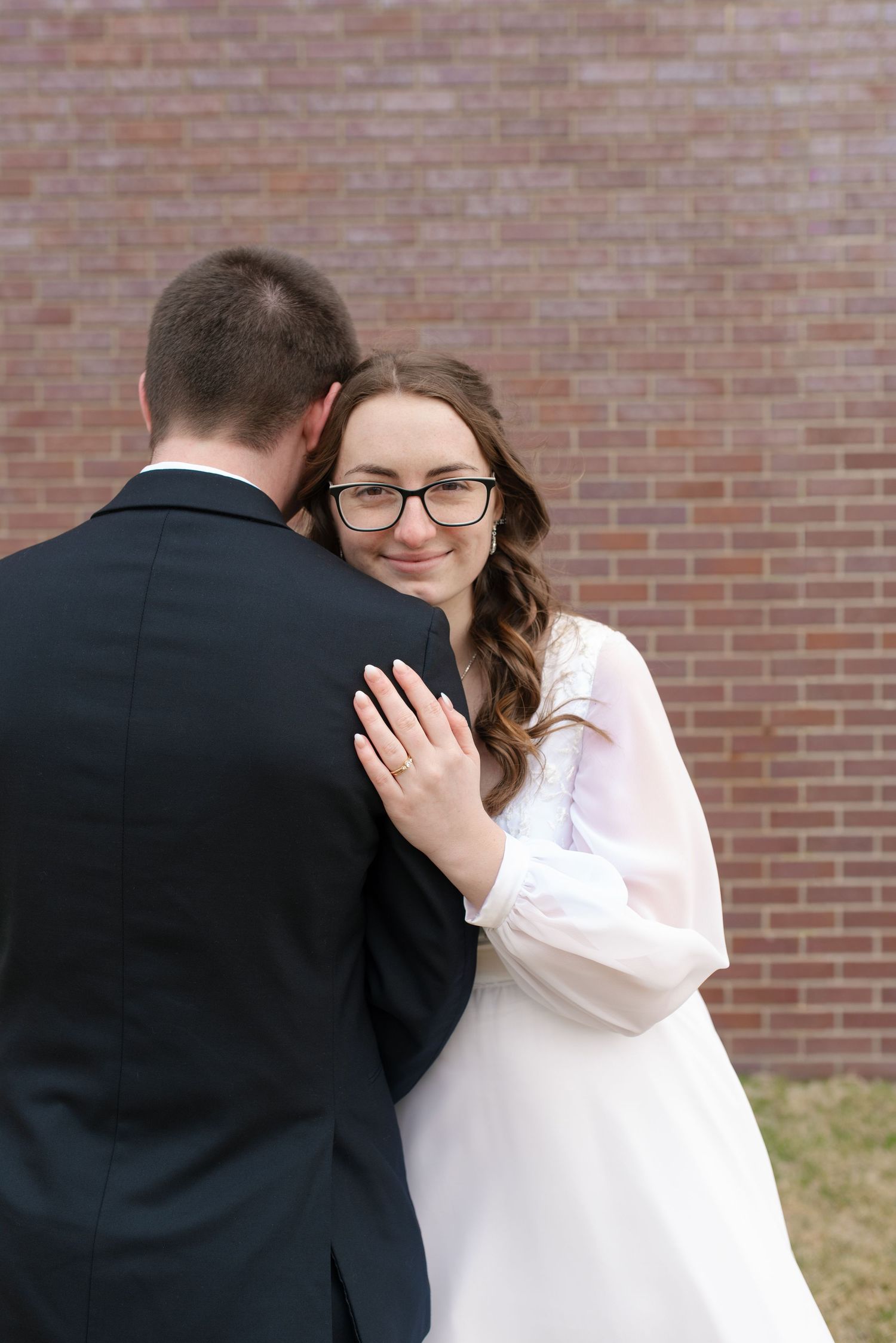 Smiling bride wearing glasses embraces her groom from behind against a red brick wall backdrop.