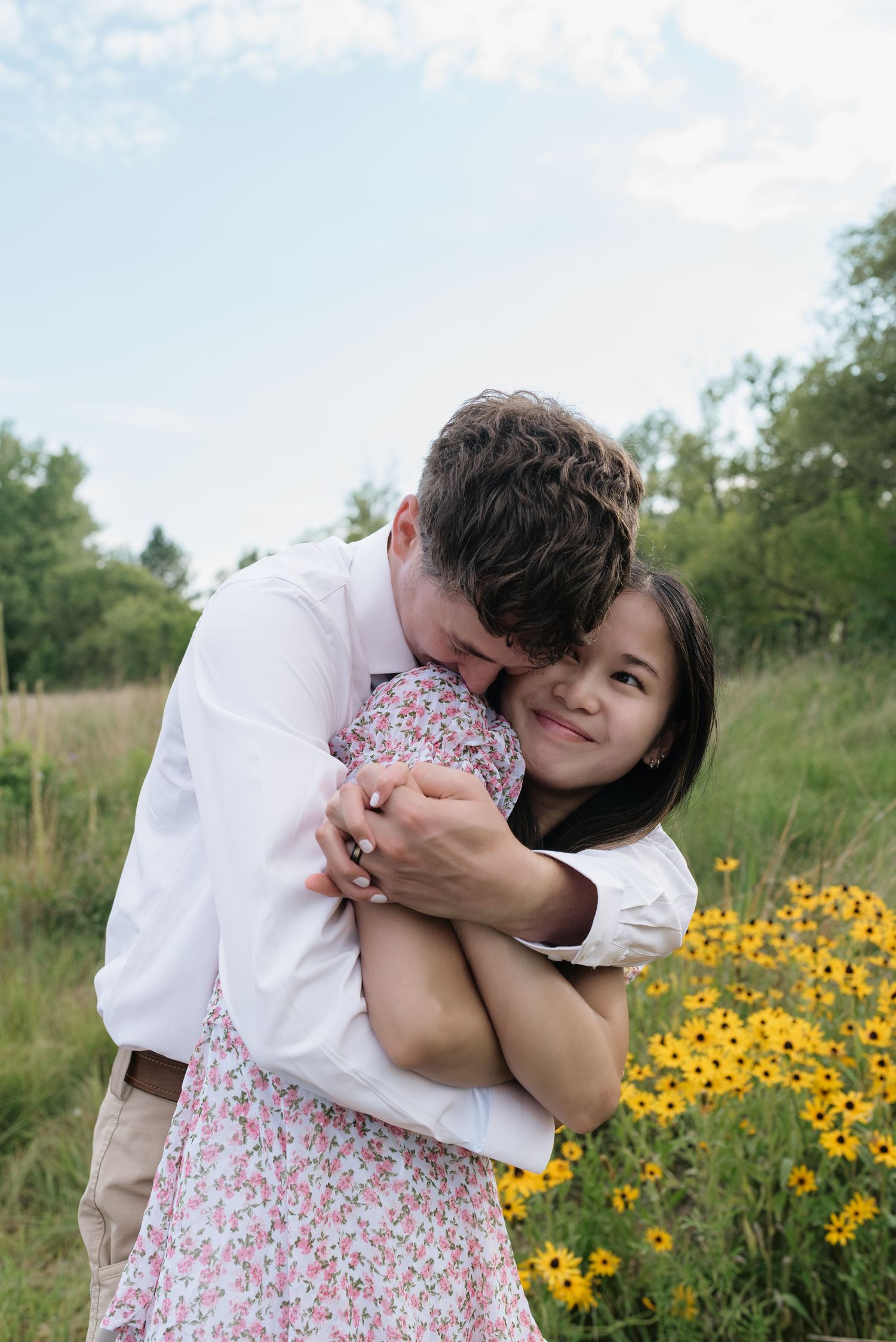 Man tenderly hugging smiling woman in floral dress near yellow wildflowers in a meadow.