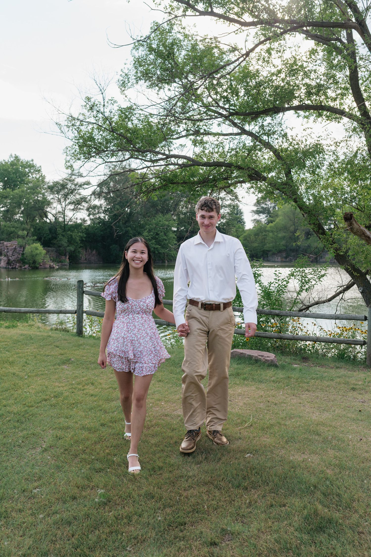 Couple holding hands walking on green grass near a pond surrounded by trees.
