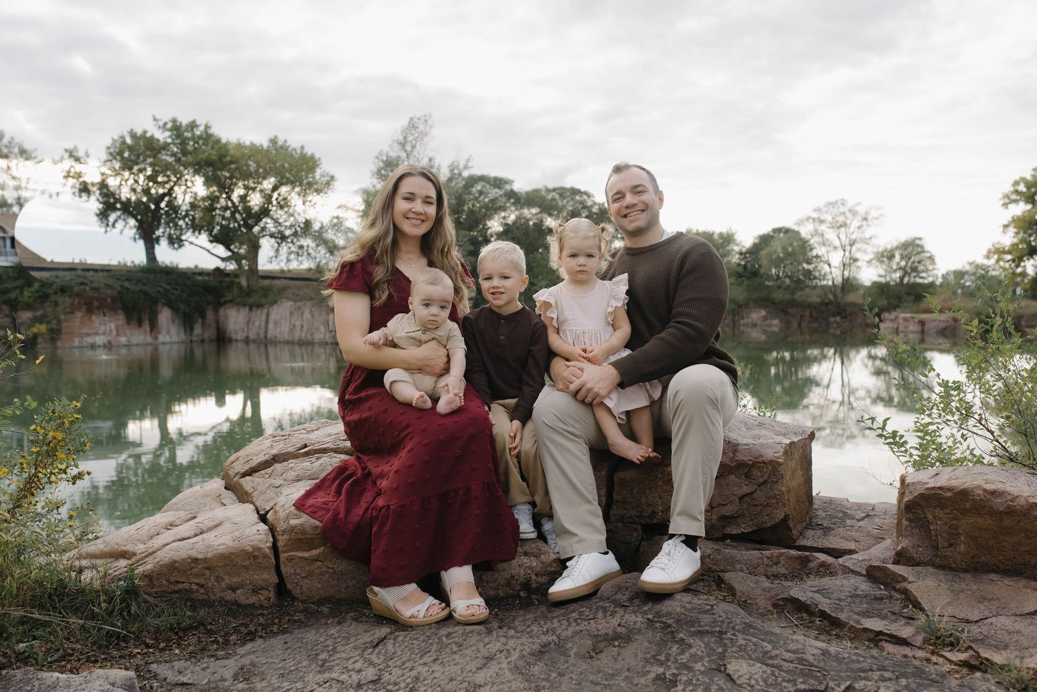 Smiling family of five posing on rocky terrain near a peaceful pond surrounded by autumn trees.