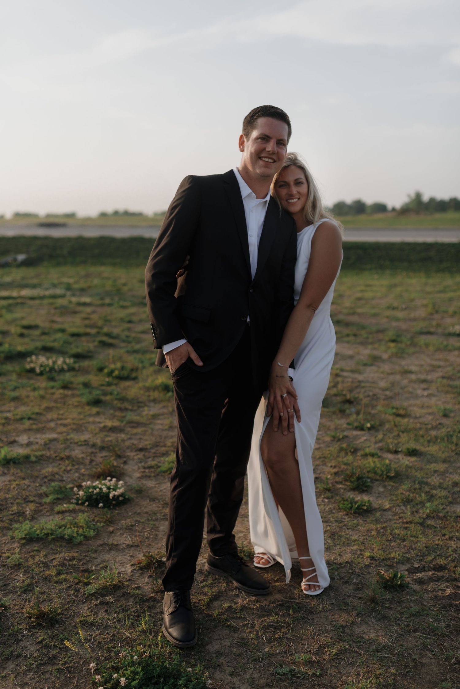 Couple posing outdoors at sunset, man in black suit and woman in white dress smiling at camera.