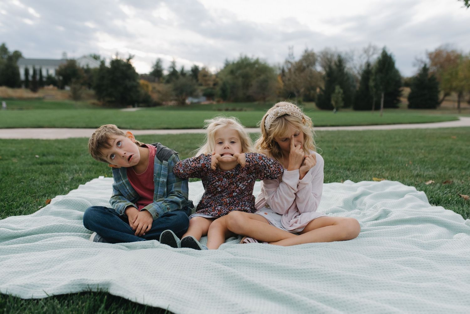 Three young children sitting on a white blanket on grass, posing playfully for a family photo outdoors.