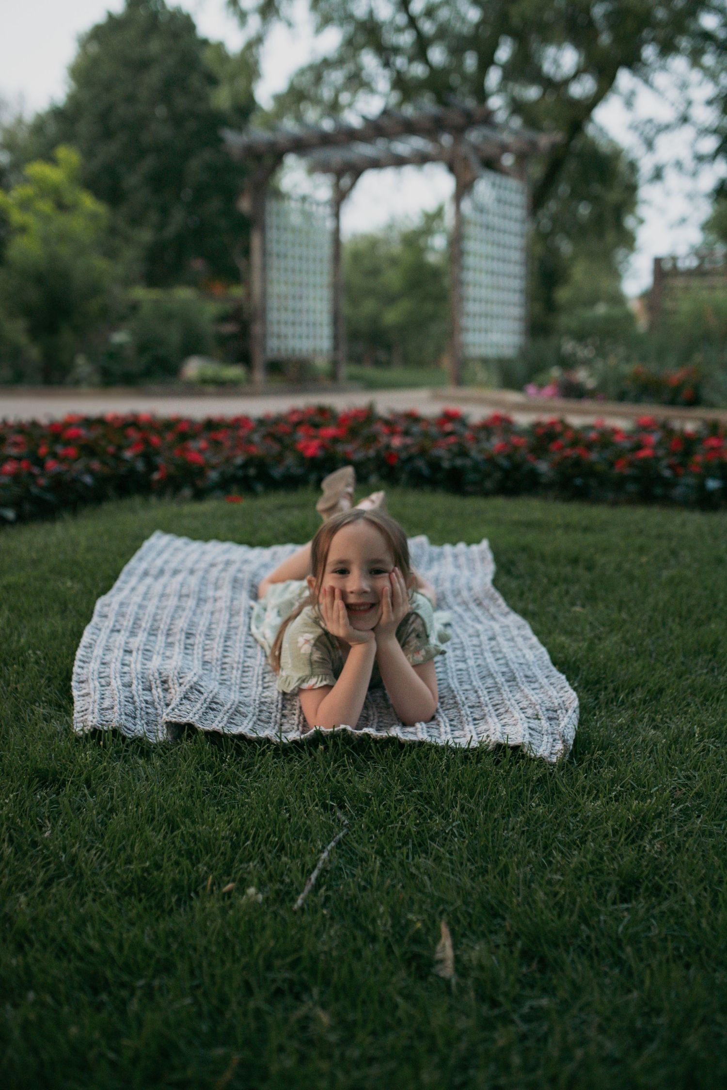 A smiling little girl lying on a crochet blanket in a garden with red flowers and an arbor.