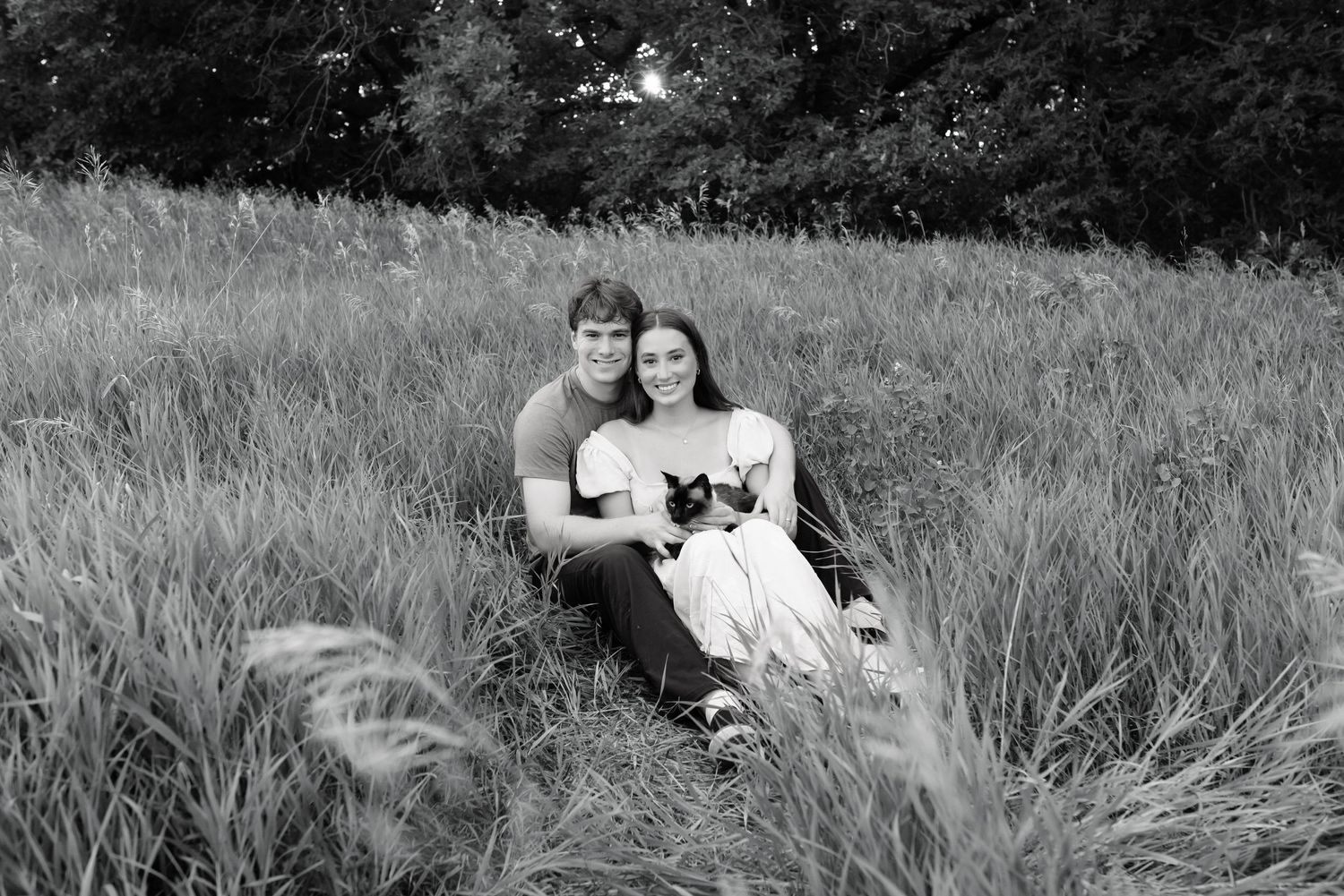 Black and white photo of a couple sitting in tall grass, smiling and holding a small cat outdoors.