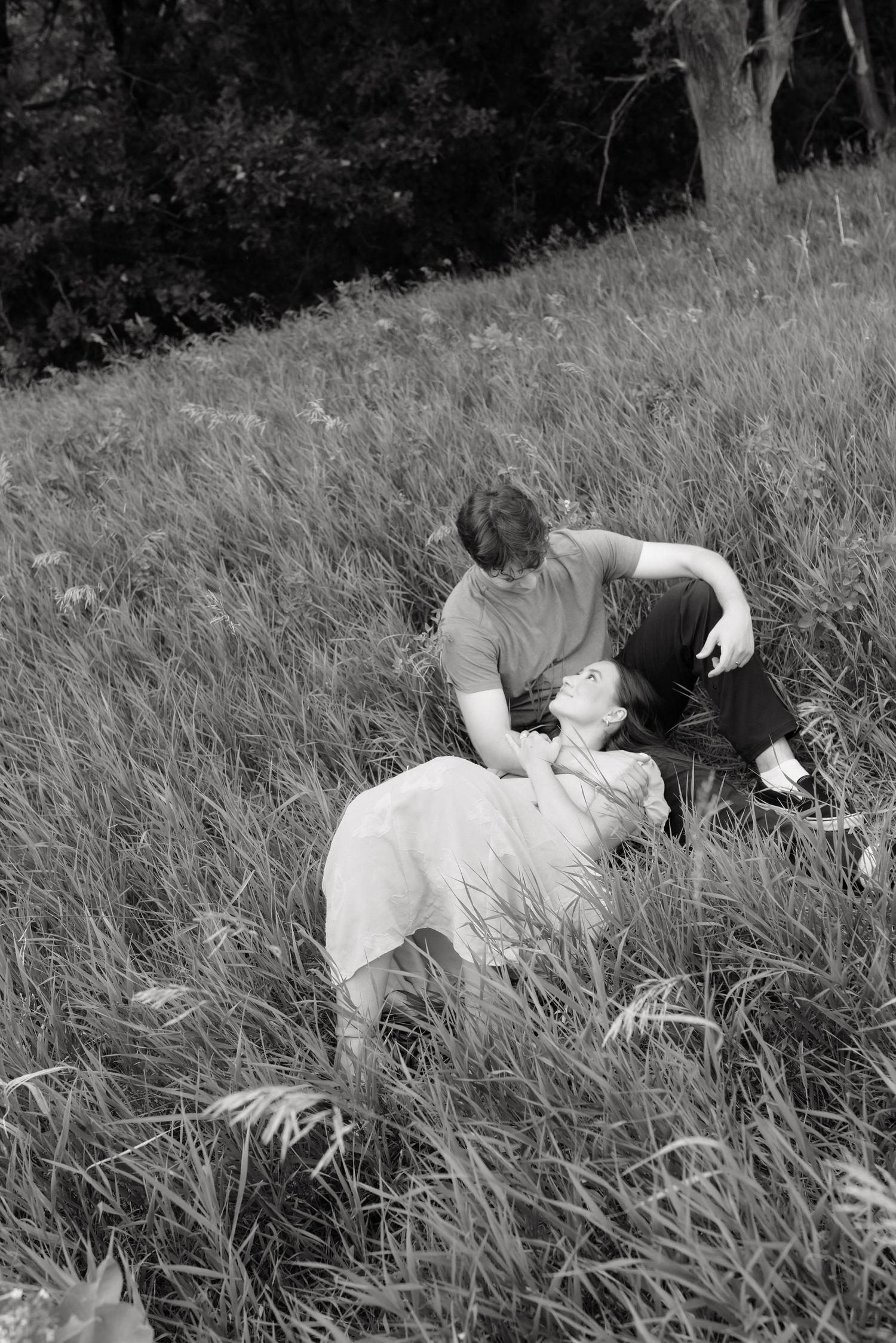 Black and white photo of a romantic couple sharing an intimate moment while lying in a lush grassy meadow.