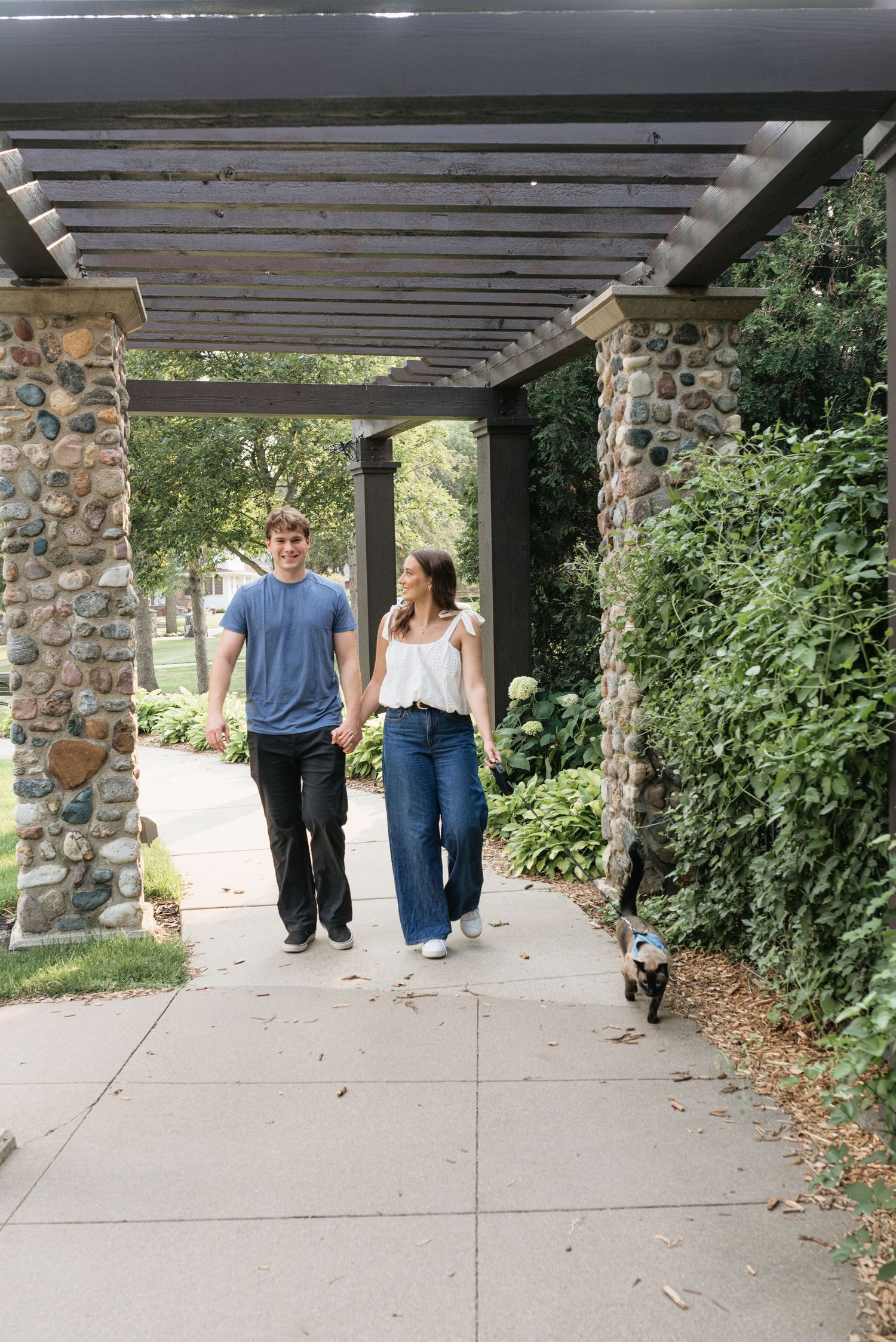 Couple holding hands walking under a stone pergola with lush greenery and a small dog nearby on a sunny day.