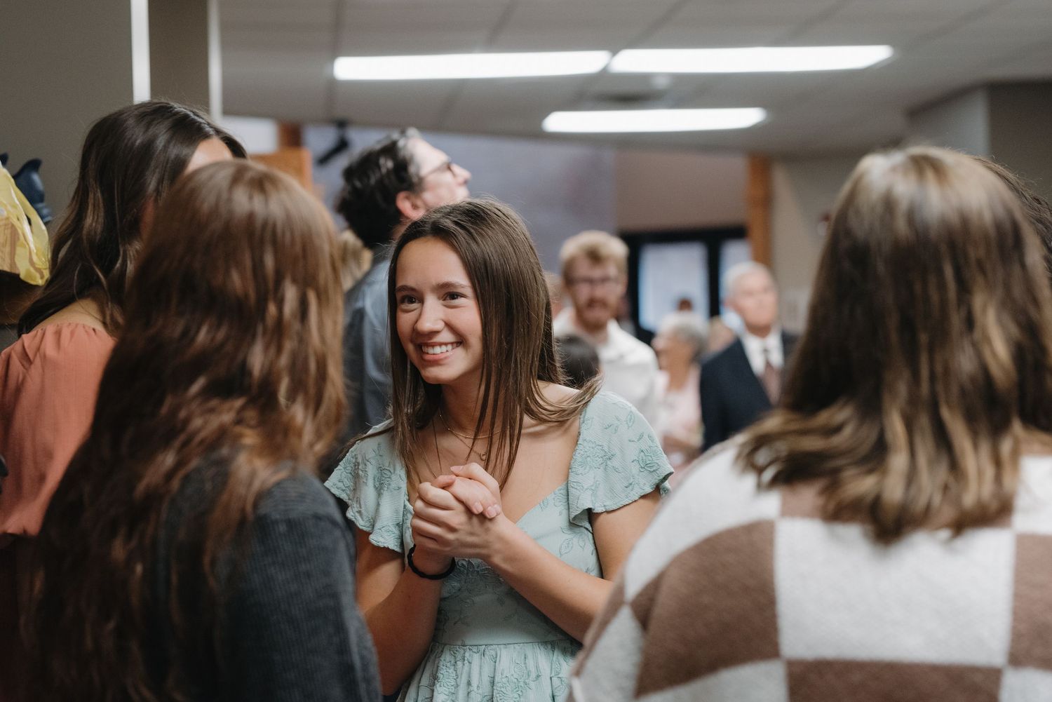 A young woman in a mint dress smiles warmly while holding hands with guests at a wedding reception.
