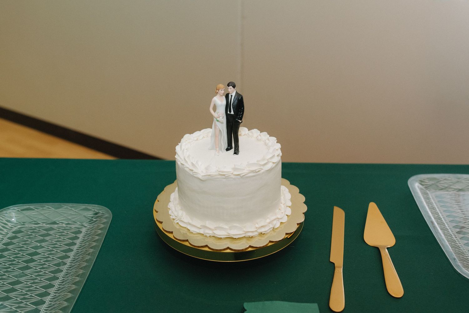 Small white wedding cake with bride and groom topper on green tablecloth with gold servers.