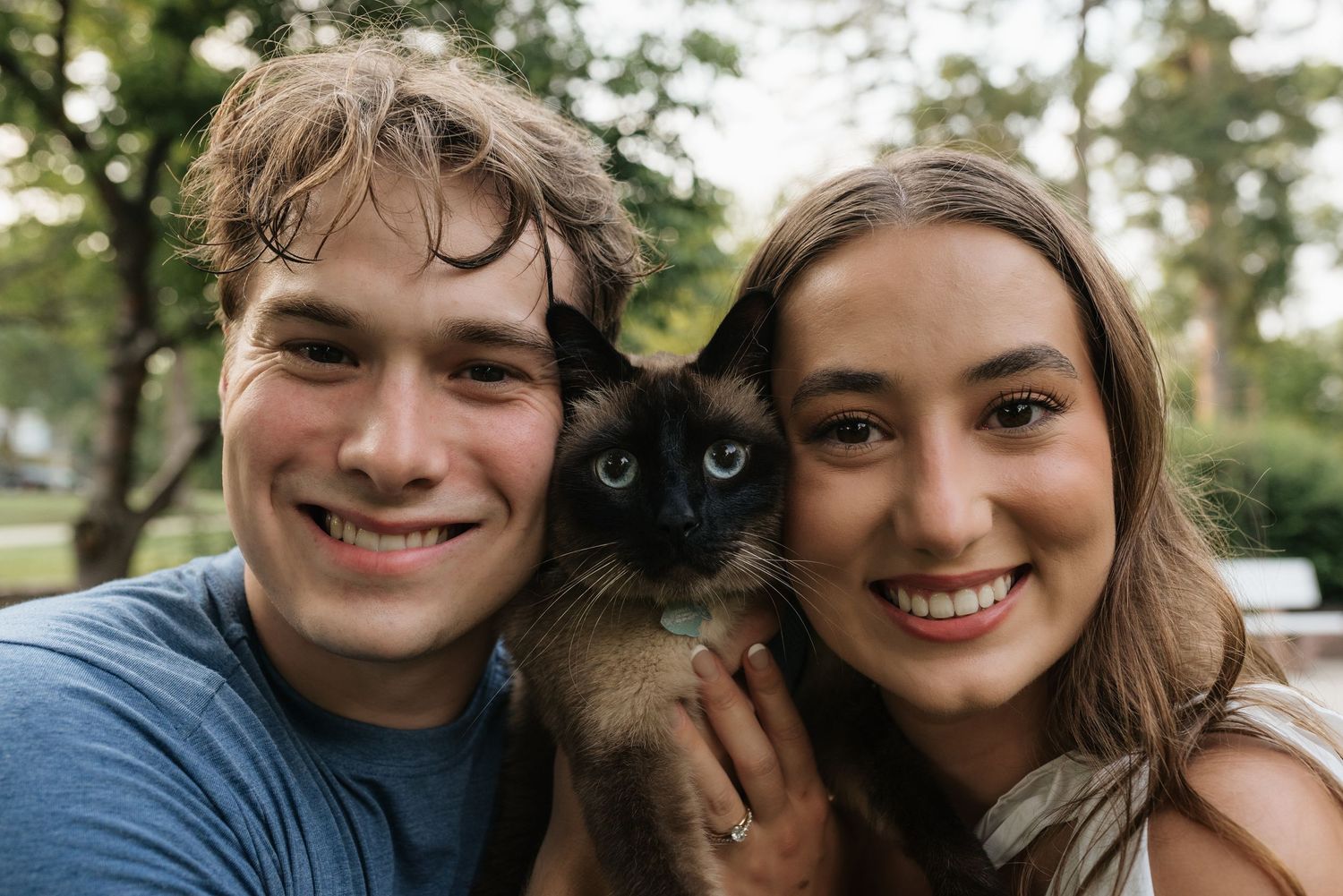 A smiling young couple takes a selfie outdoors while holding a Siamese cat in a lush green park setting.