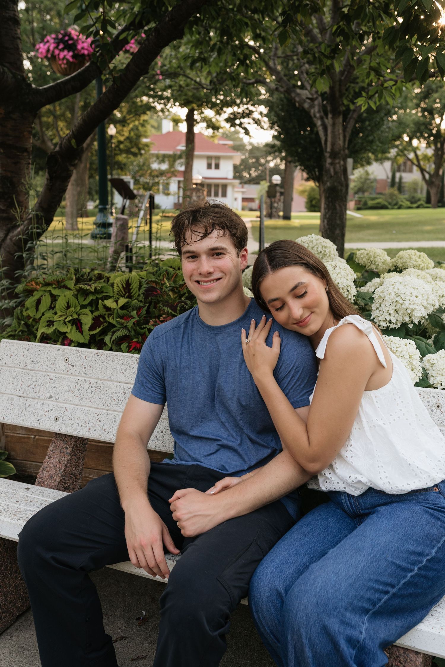 A smiling couple sits on a white bench surrounded by lush greenery and white hydrangeas in a park setting.