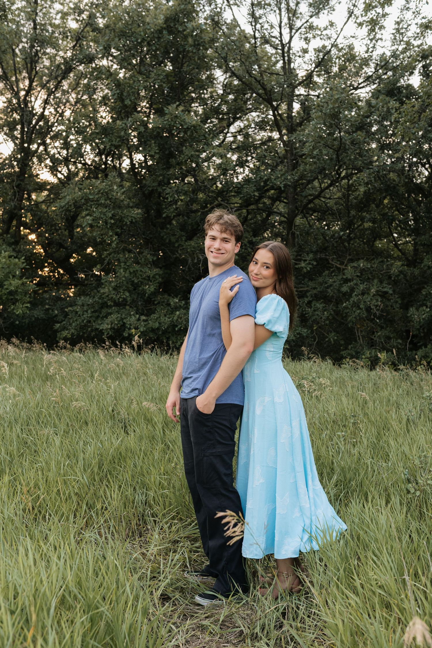 A young couple poses in a lush green field, the woman in a light blue dress leaning on the man in a blue shirt.