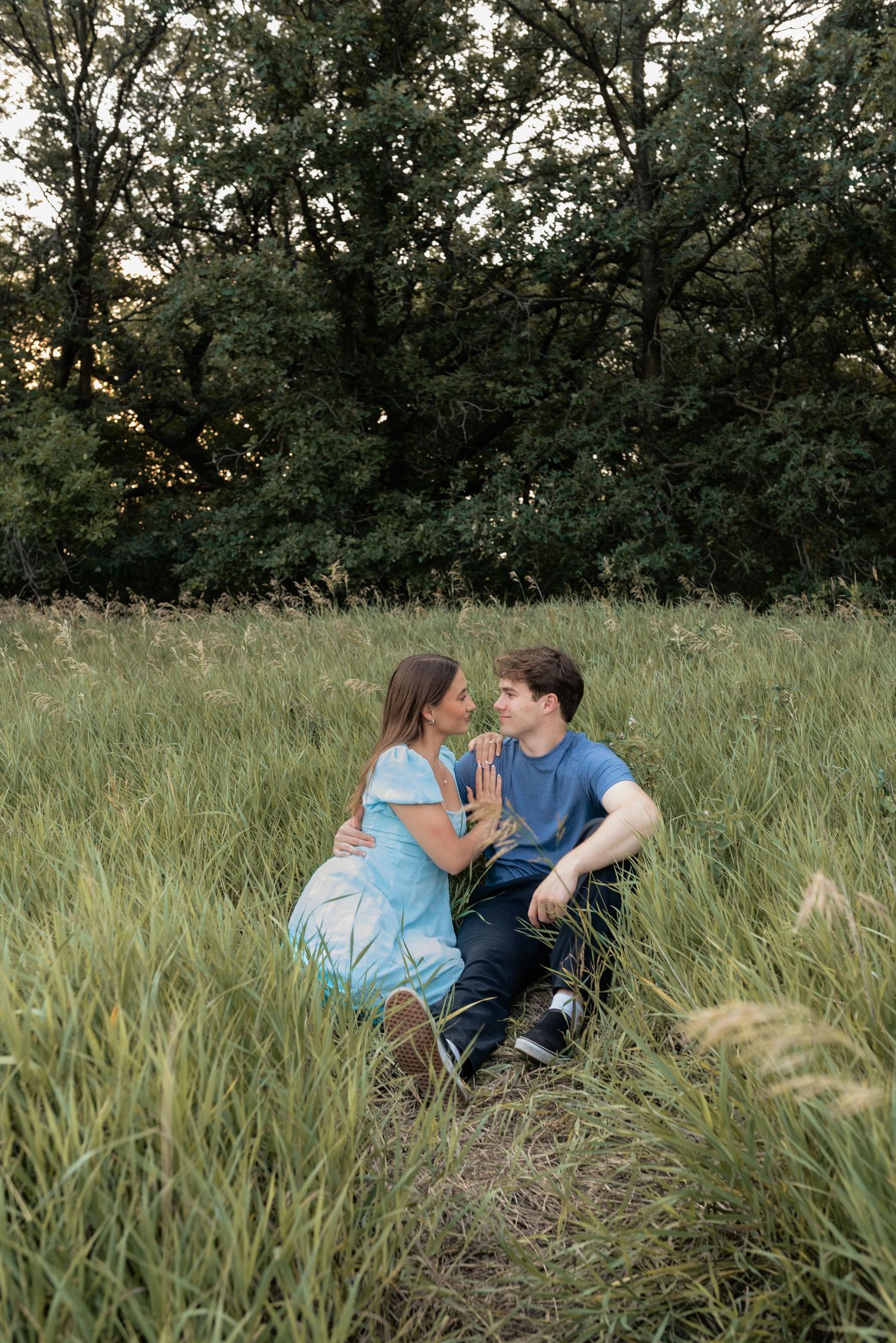 A couple sits together in a lush green meadow, sharing a tender moment surrounded by tall grass and trees.