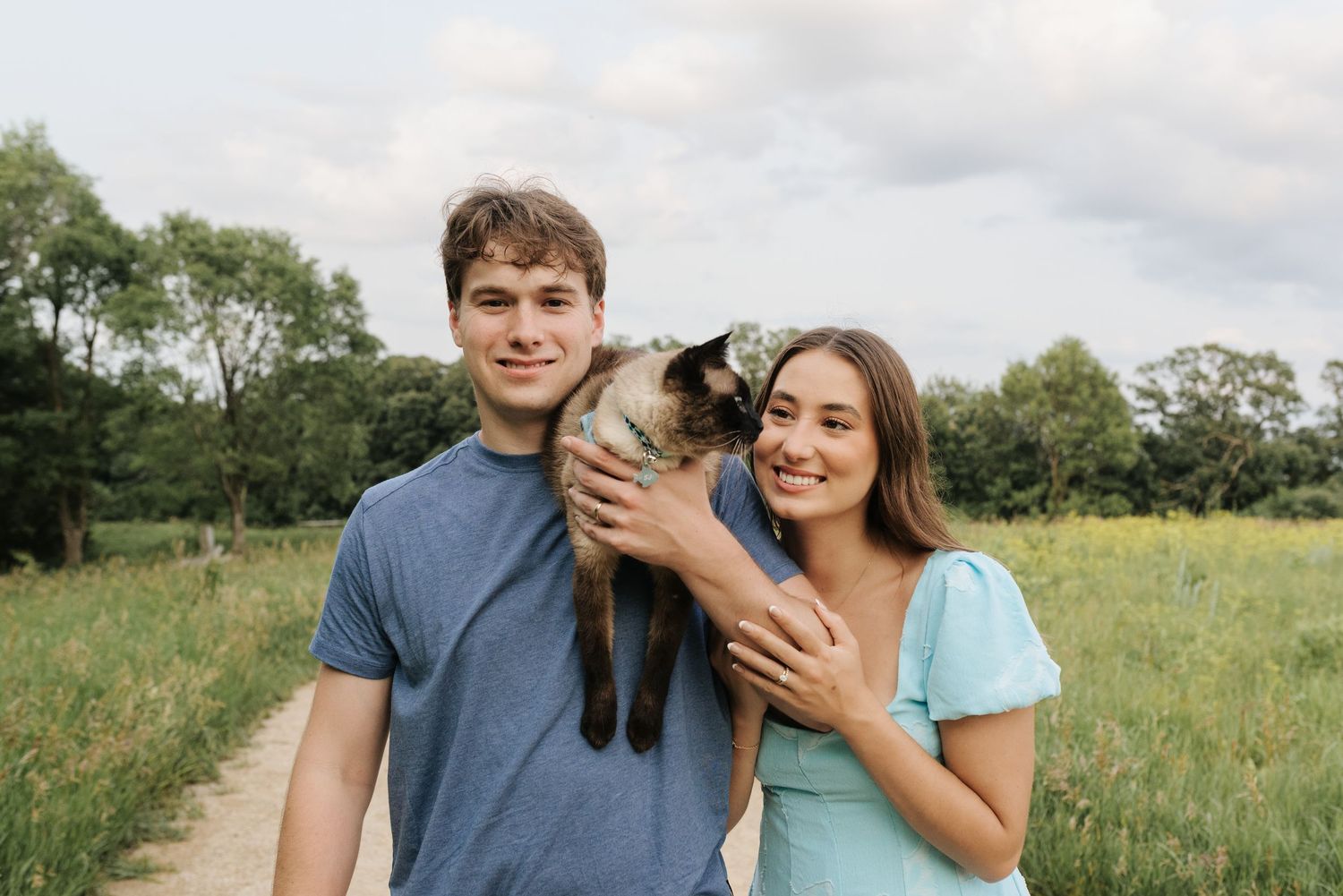 Young couple smiling outdoors holding a Siamese cat on a scenic green field path.