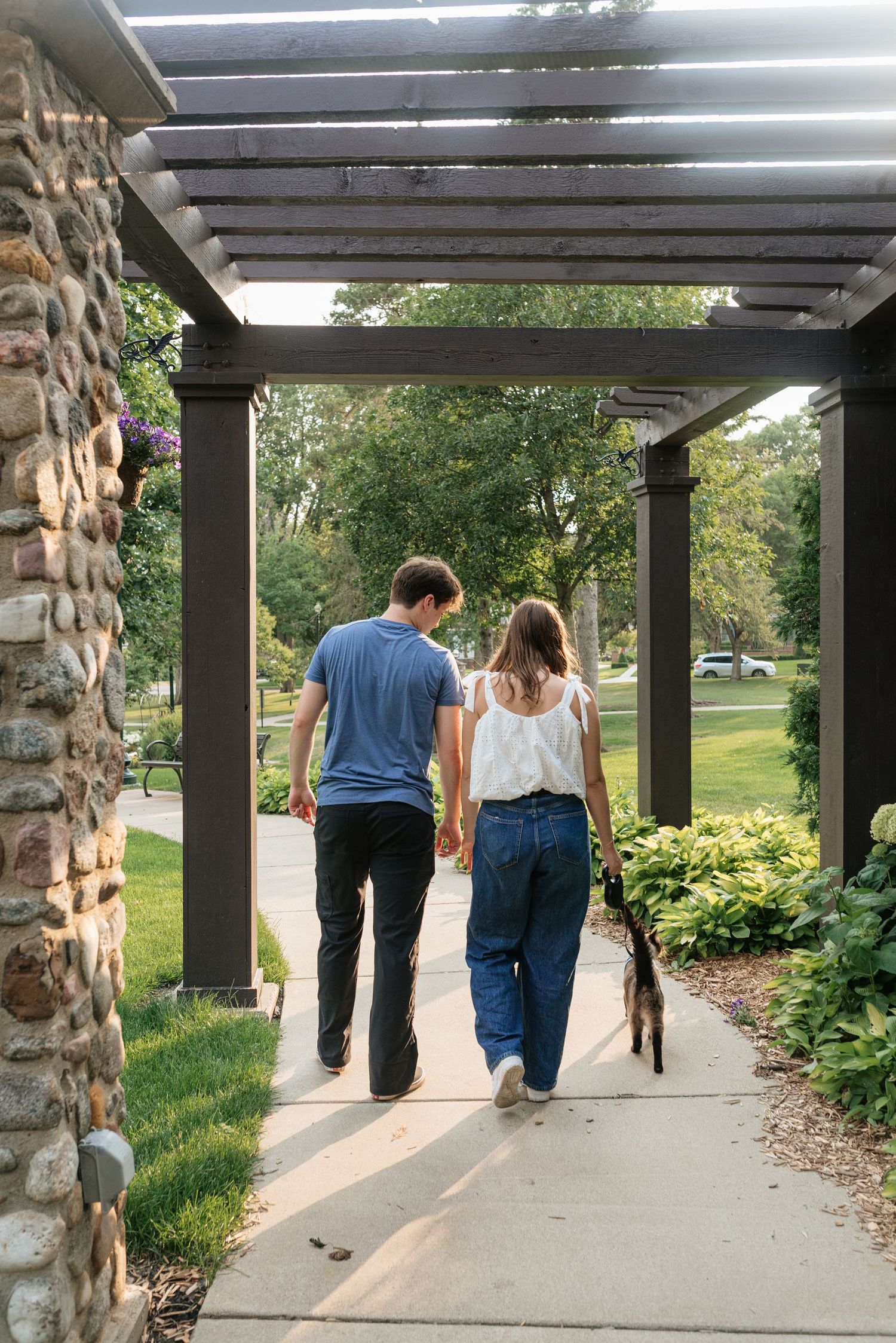 Couple walking dog under pergola on garden path surrounded by lush greenery and stone pillars.