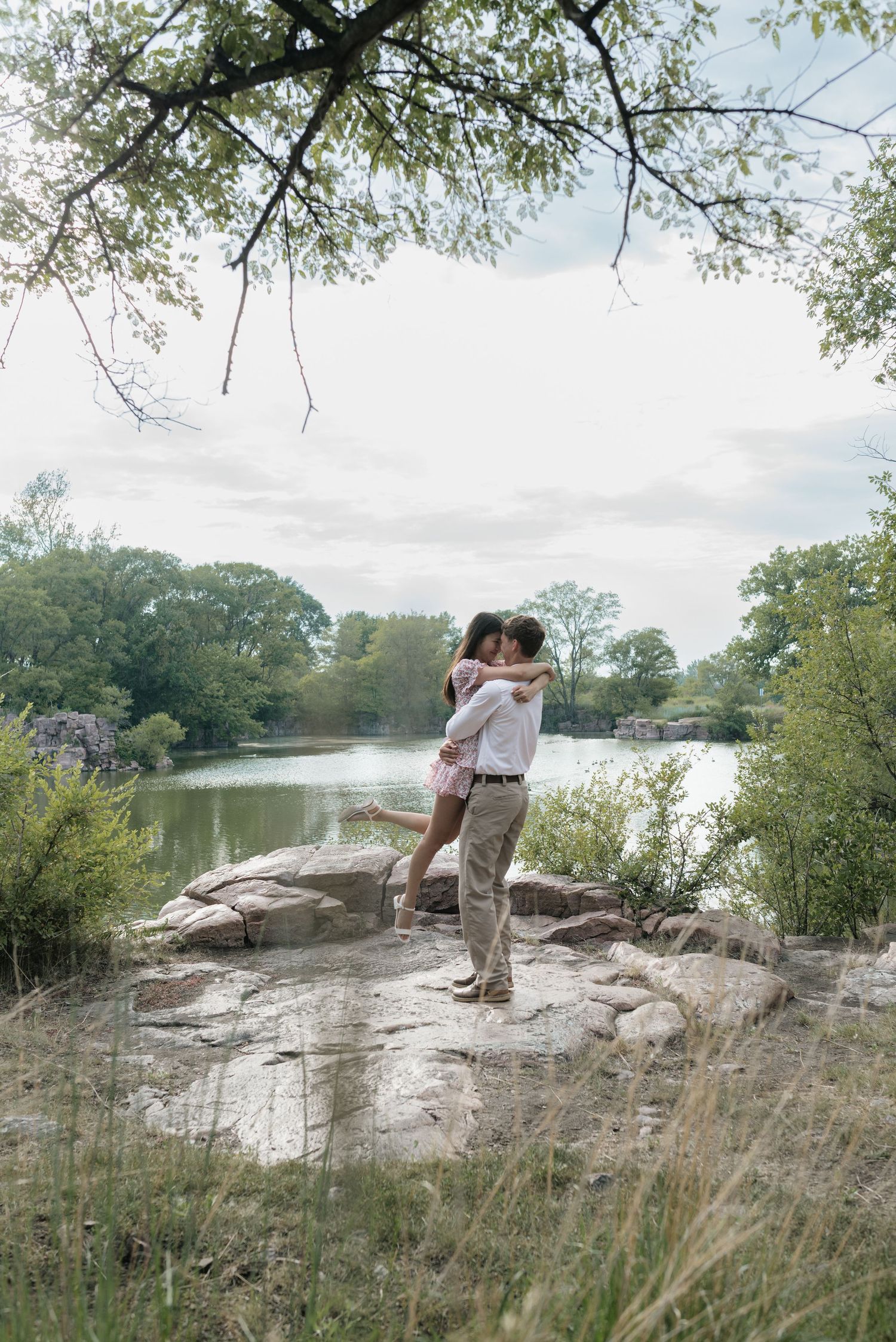 Couple embracing and kissing on rocks by a serene lake surrounded by lush green trees.