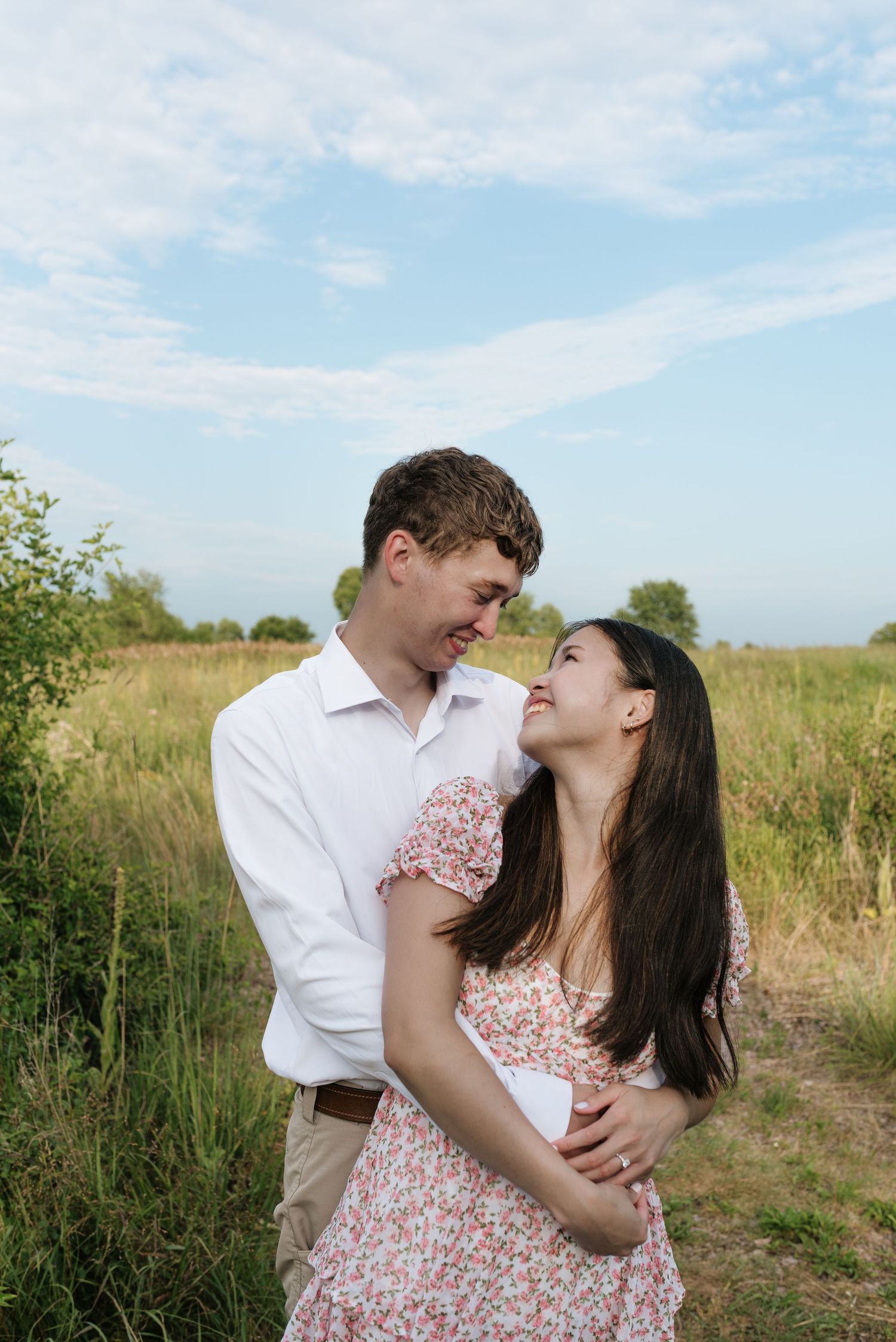 A couple gazes lovingly at each other while embracing in an open grassy field under a blue sky.