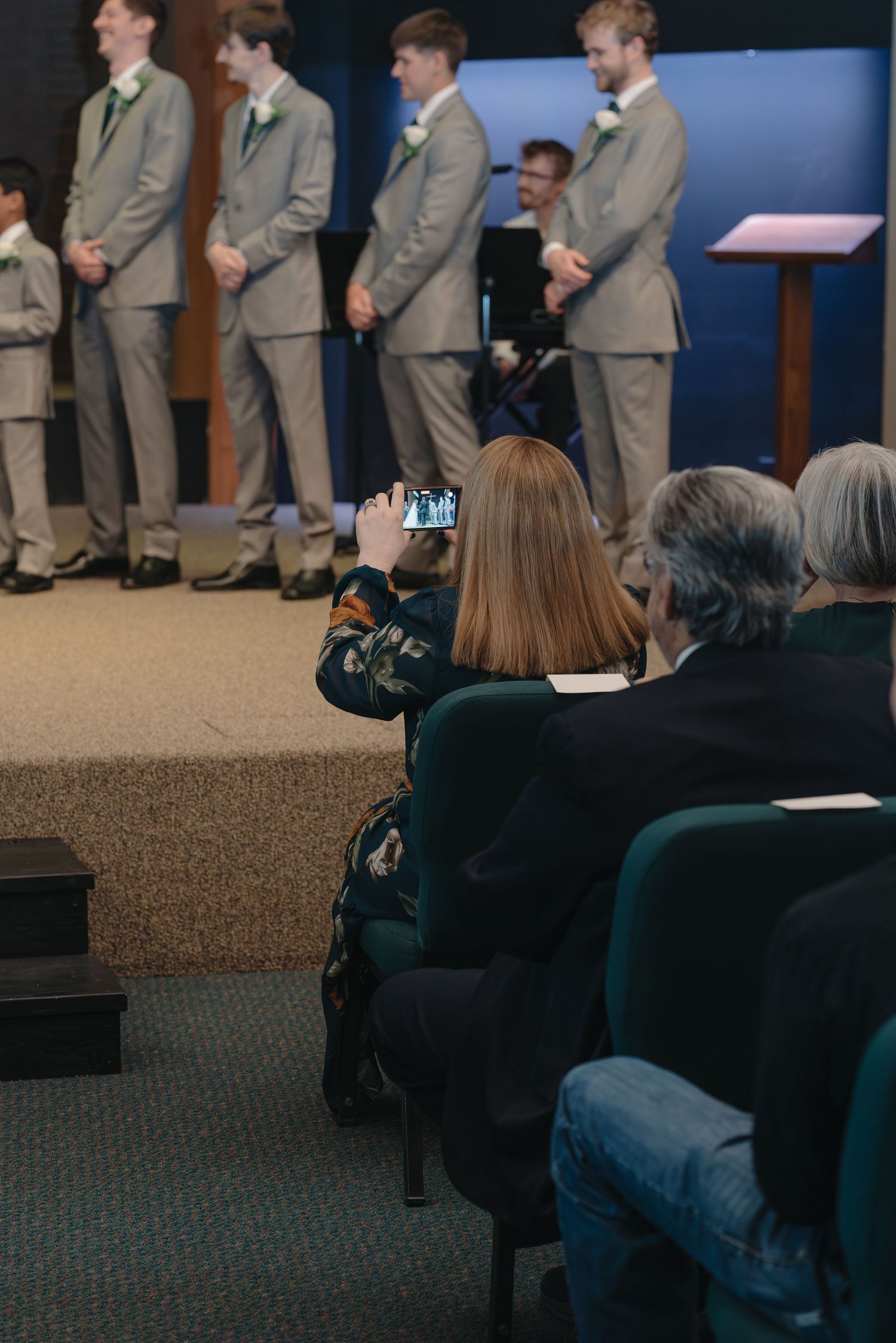 Wedding guests photograph groomsmen in tan suits standing at altar during indoor ceremony.