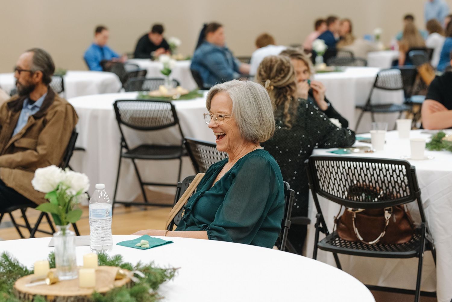 Elegantly dressed woman laughing joyfully at wedding reception seated at white floral table.