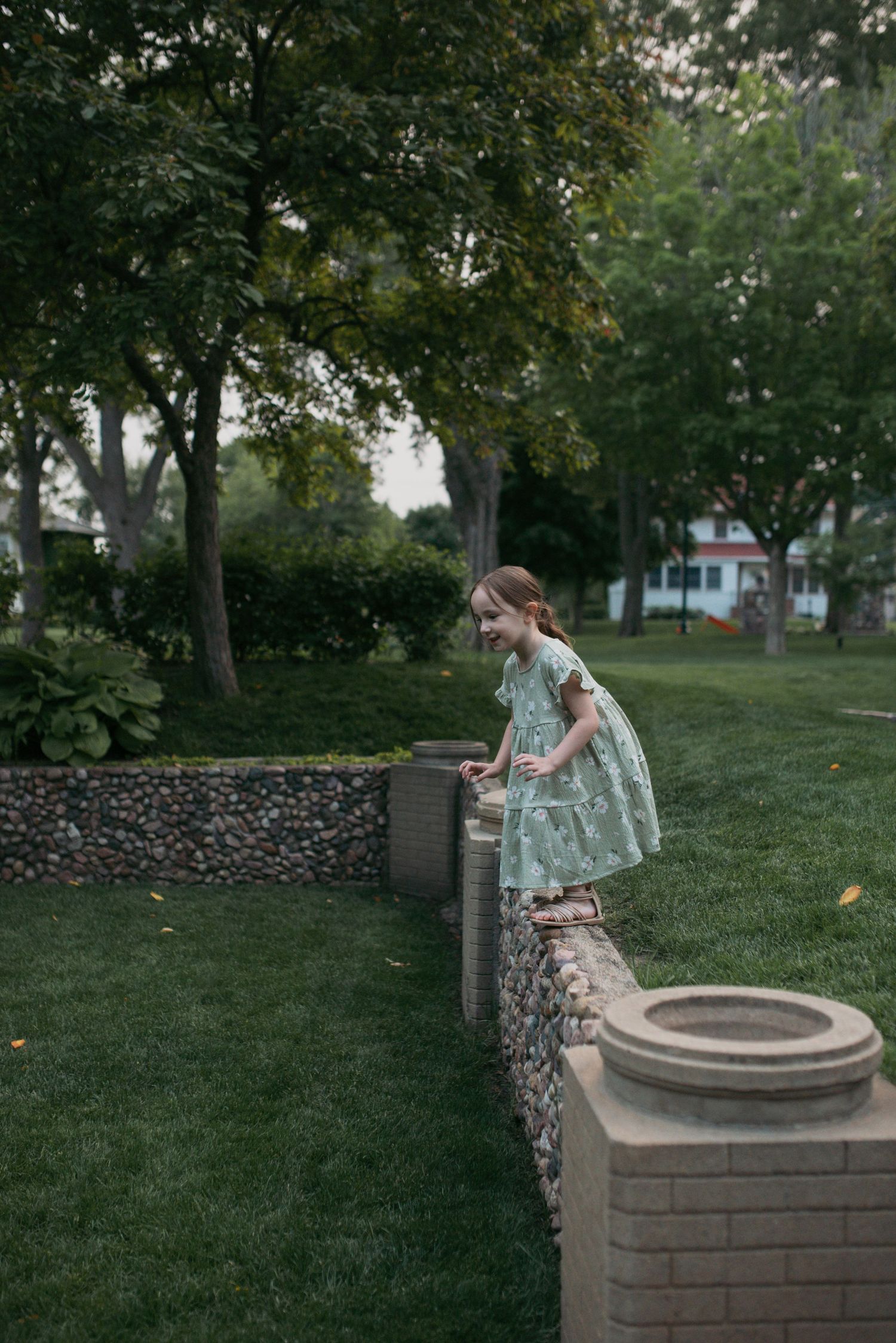 A young girl in a mint floral dress balancing on a stone garden wall in a lush green park.