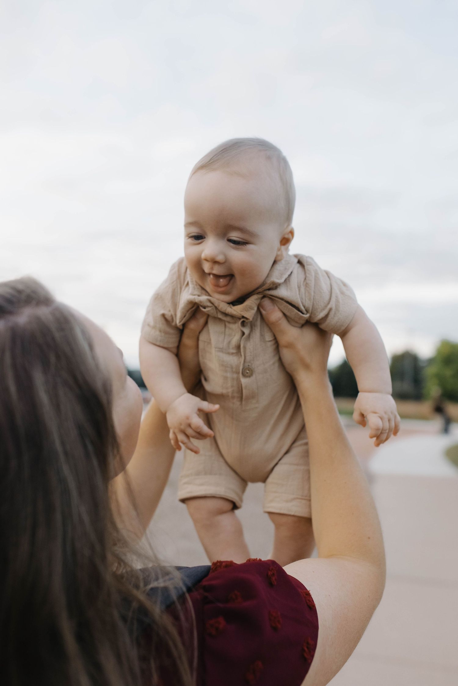 Smiling baby in beige romper lifted joyfully by parent on an outdoor path surrounded by greenery.