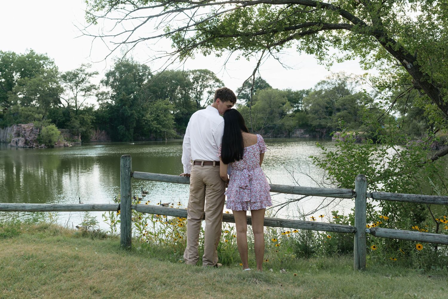 Couple standing by a wooden fence overlooking a serene lake surrounded by lush green trees.