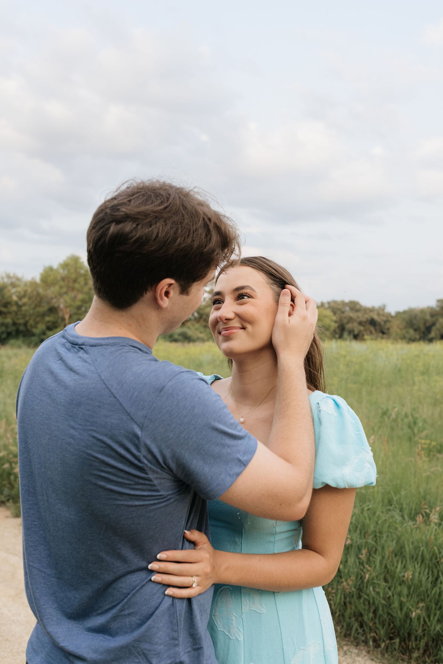 A smiling young woman in a teal dress embraces a man in a blue t-shirt in a green field.