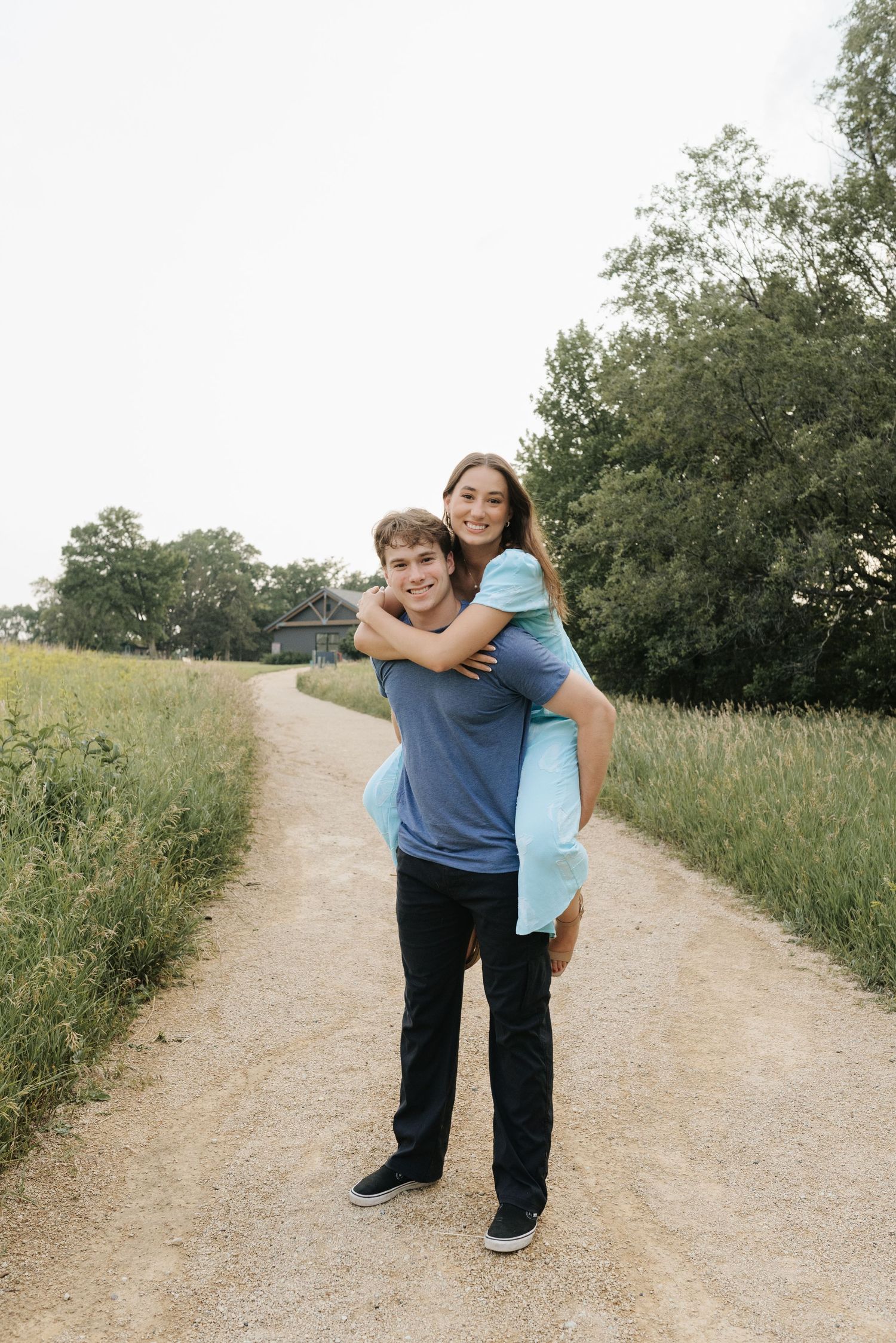 Young couple sharing a piggyback ride on a scenic dirt path surrounded by green fields and trees.
