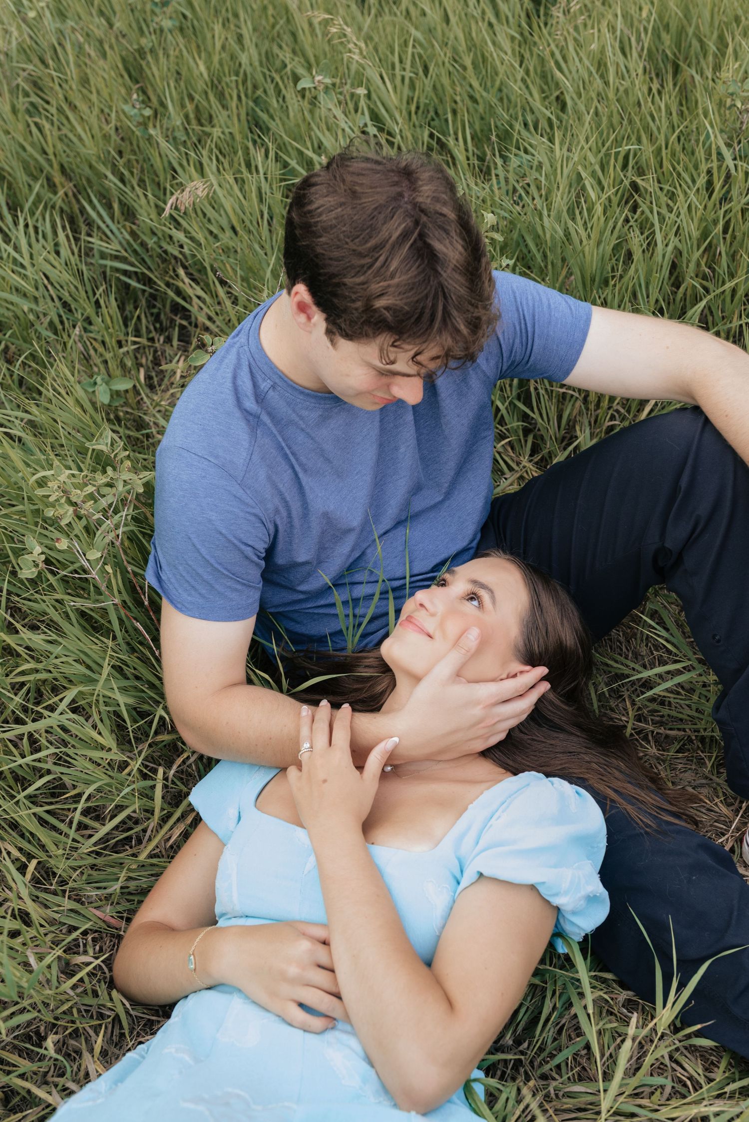 A couple shares a romantic moment in a lush green field, with the woman lying in the man's lap smiling up at him.