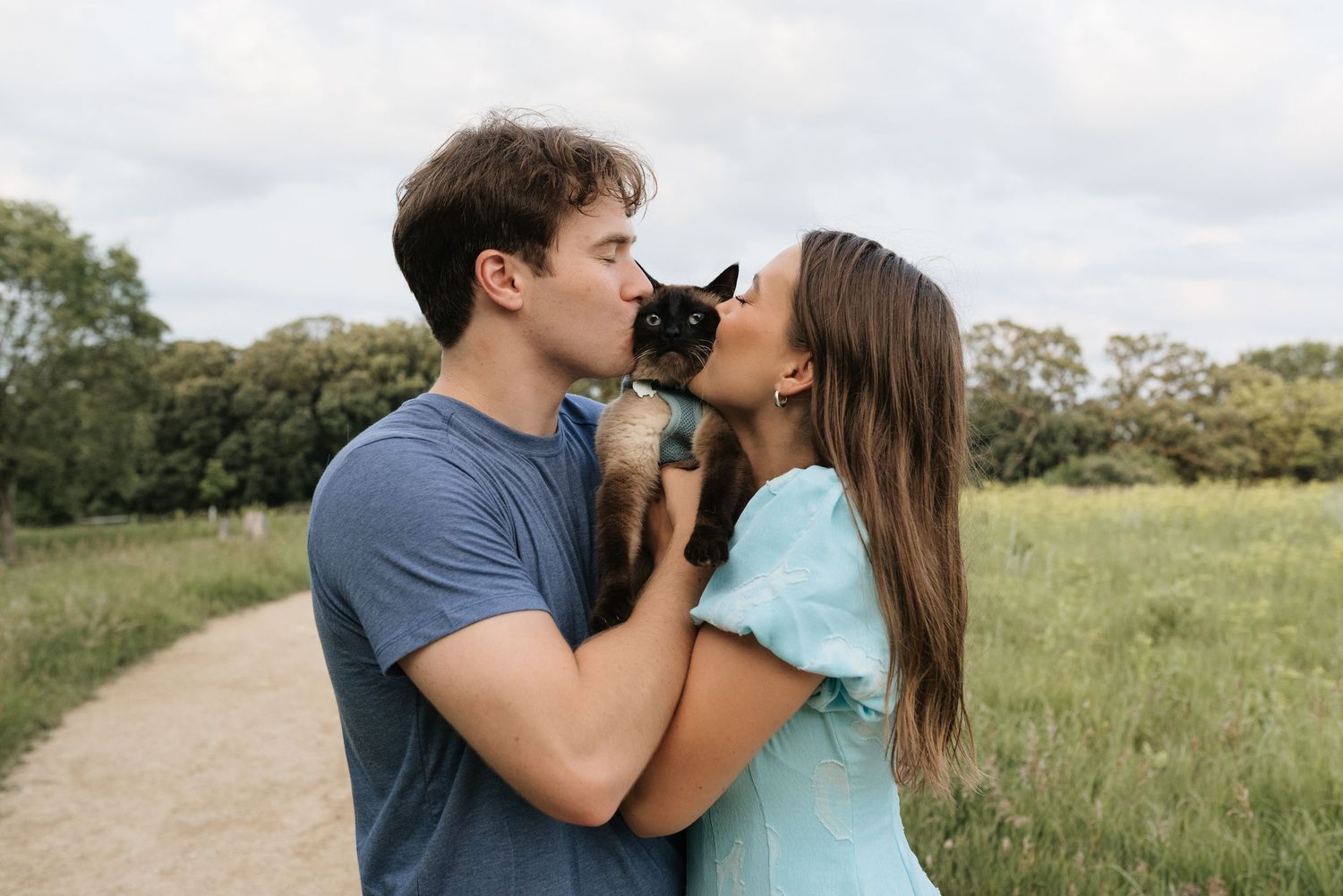 A young couple shares a kiss over a small black cat outdoors on a scenic park path surrounded by greenery.