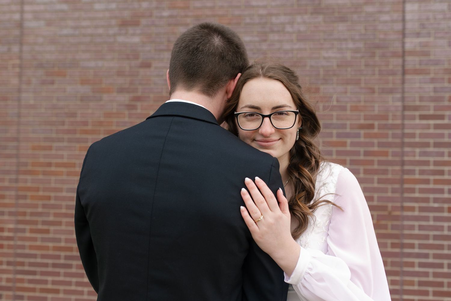Bride in white dress embraces groom, smiling warmly while showing wedding ring on her hand.