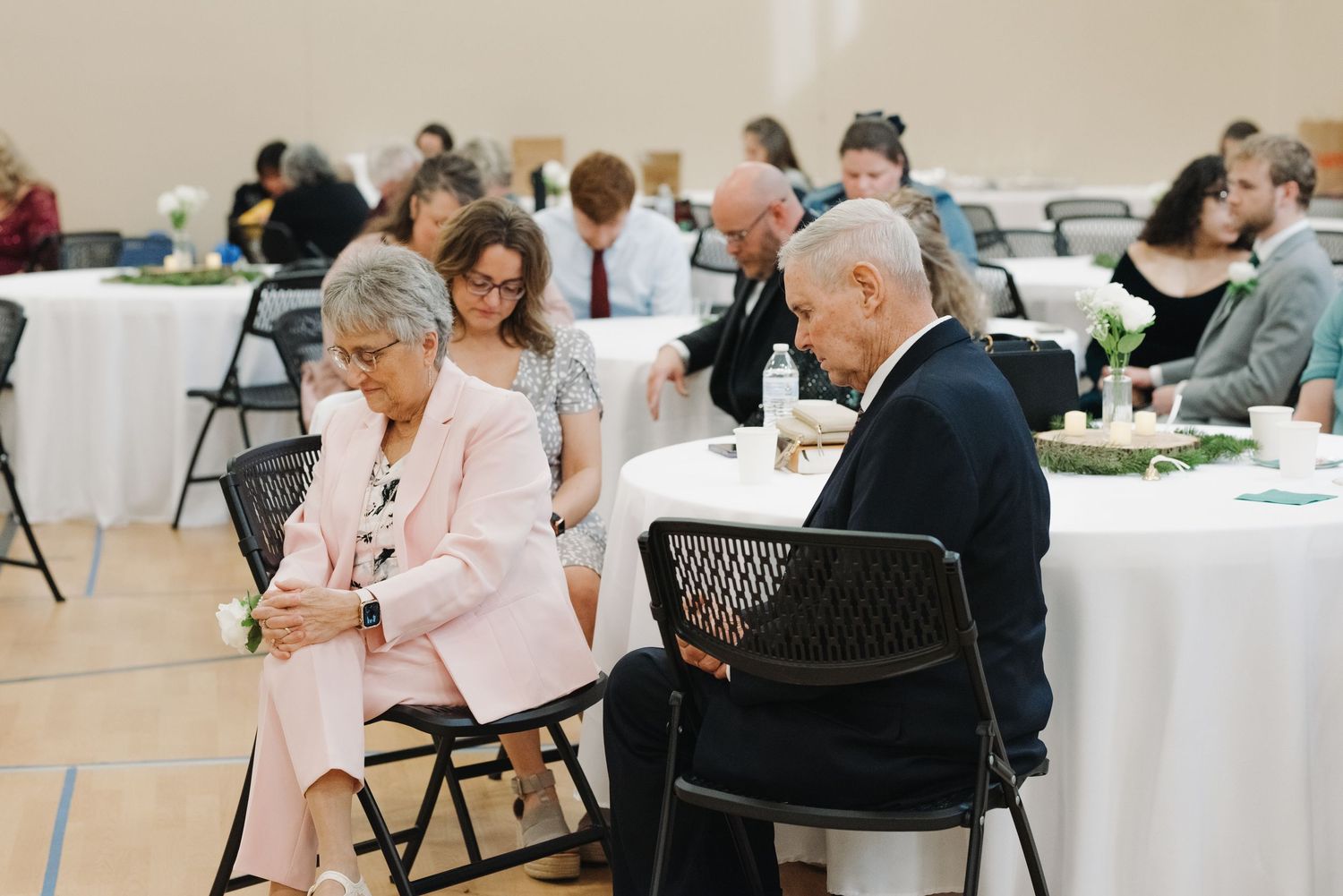Wedding guests seated at round tables with white linens, bowing heads in prayer at reception.
