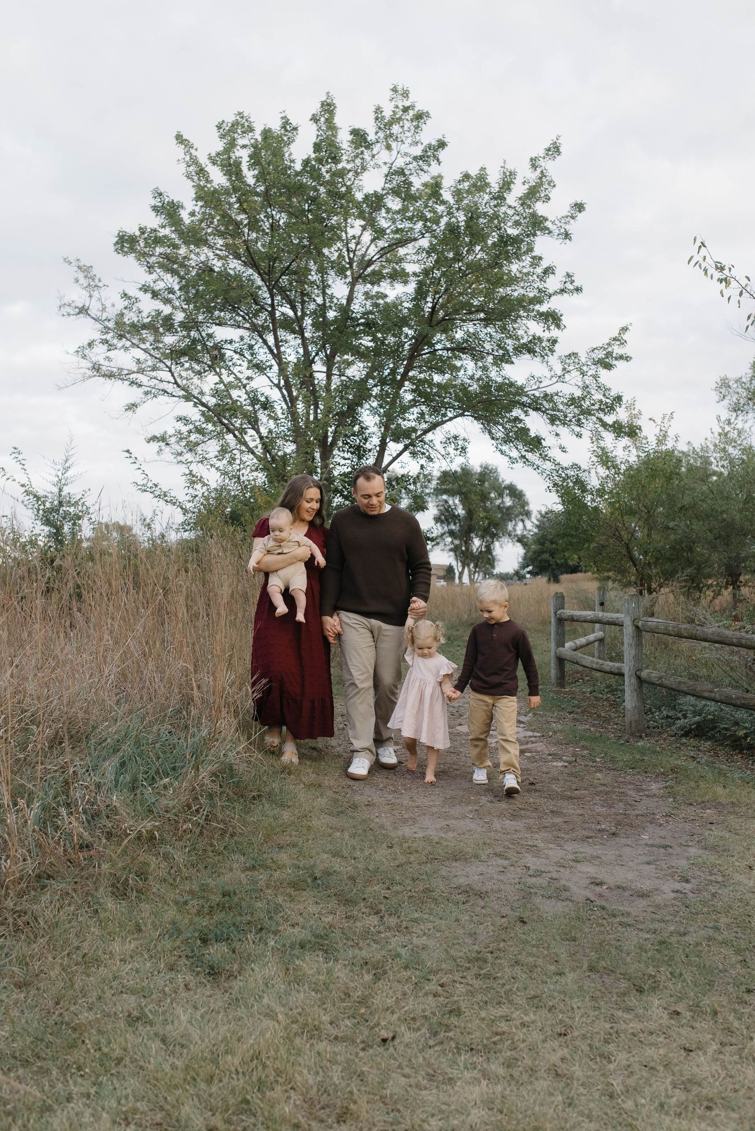 Happy family of four walking along a dirt path surrounded by tall grass and a large tree.