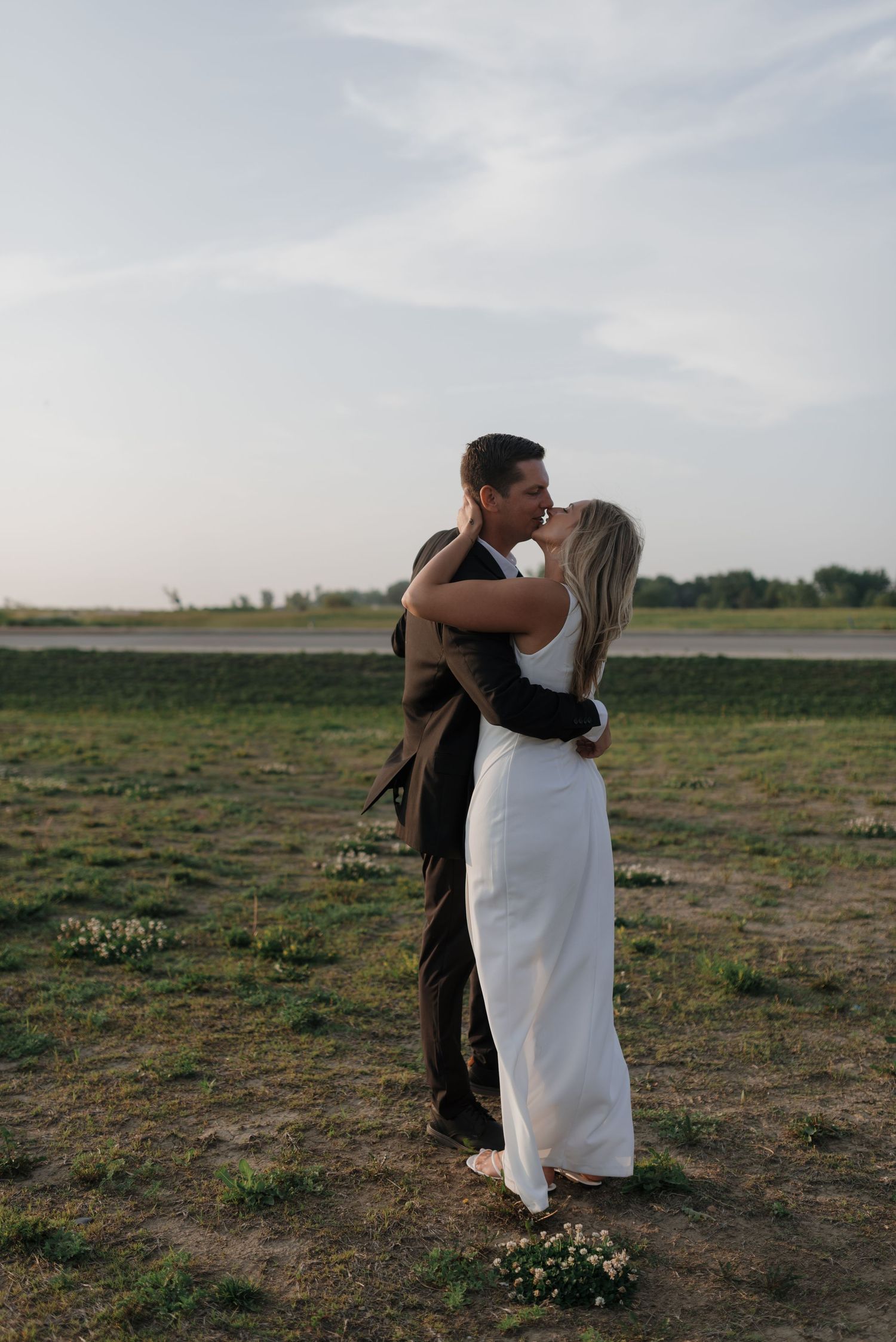 Bride and groom share a romantic kiss outdoors in green field during wedding photo session.