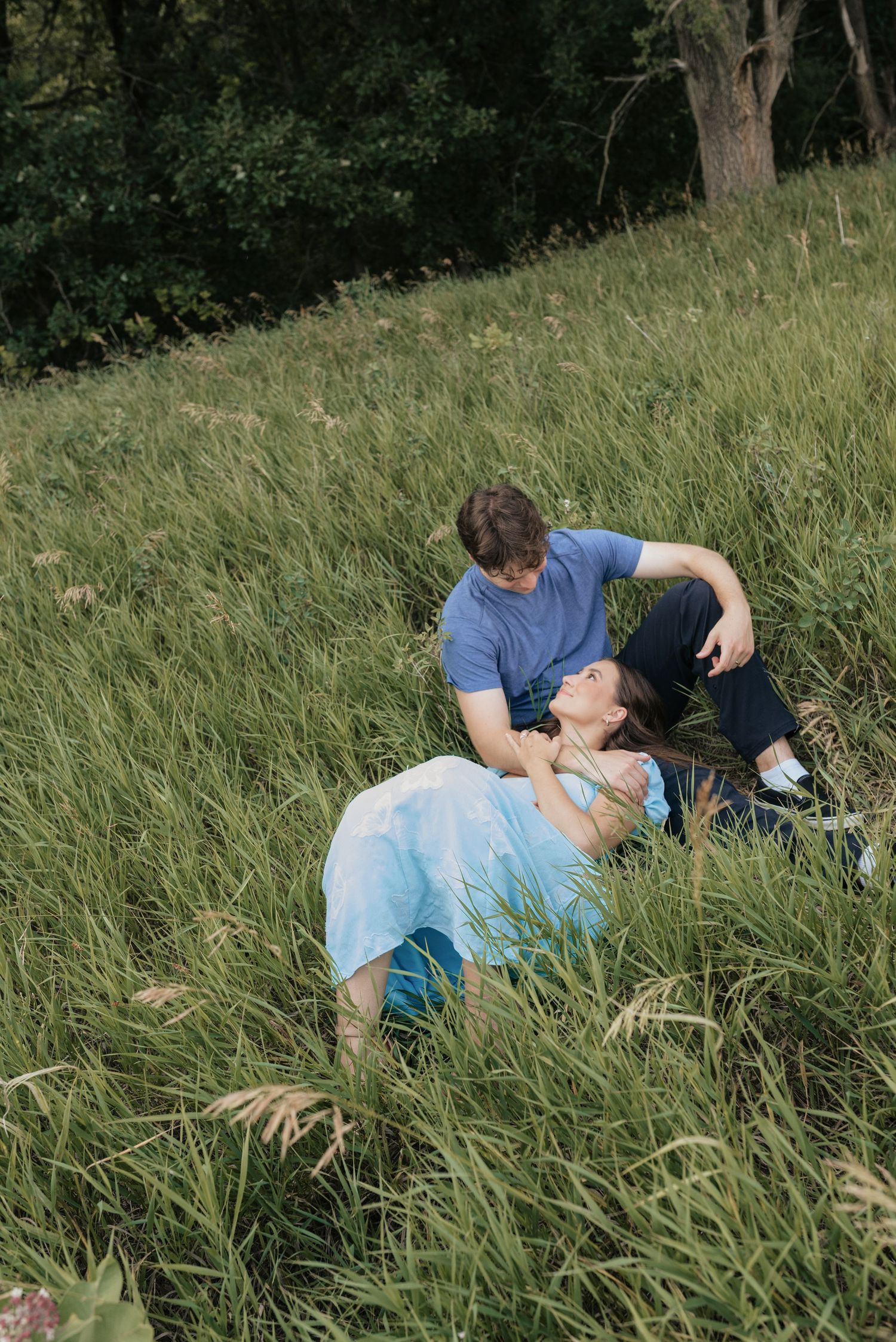 A couple relaxes in tall green grass, the woman lying back laughing as the man sits beside her outdoors.