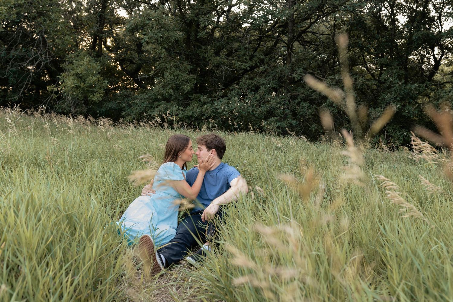 Couple sharing a romantic kiss while sitting in tall green grass field with lush trees in background.