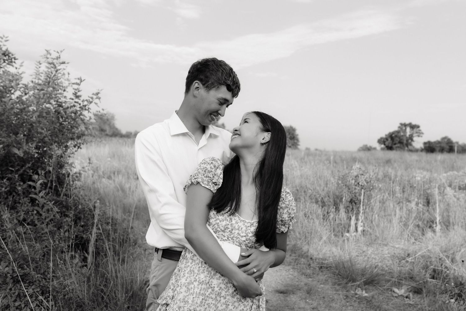 Black and white photo of a smiling couple embracing in an open field under a soft cloudy sky.