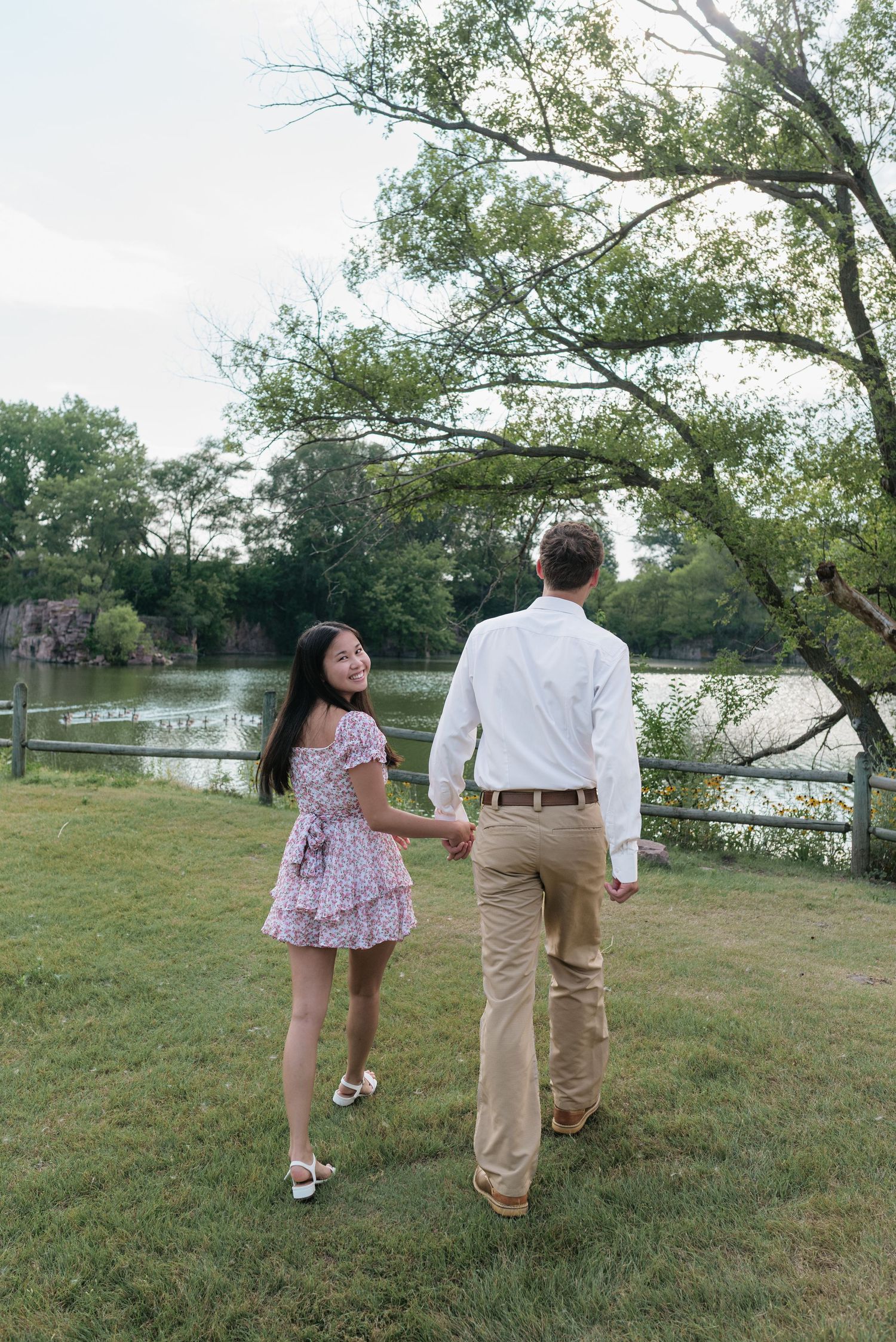 Couple holding hands walking toward a scenic pond surrounded by lush green trees on a warm summer day.