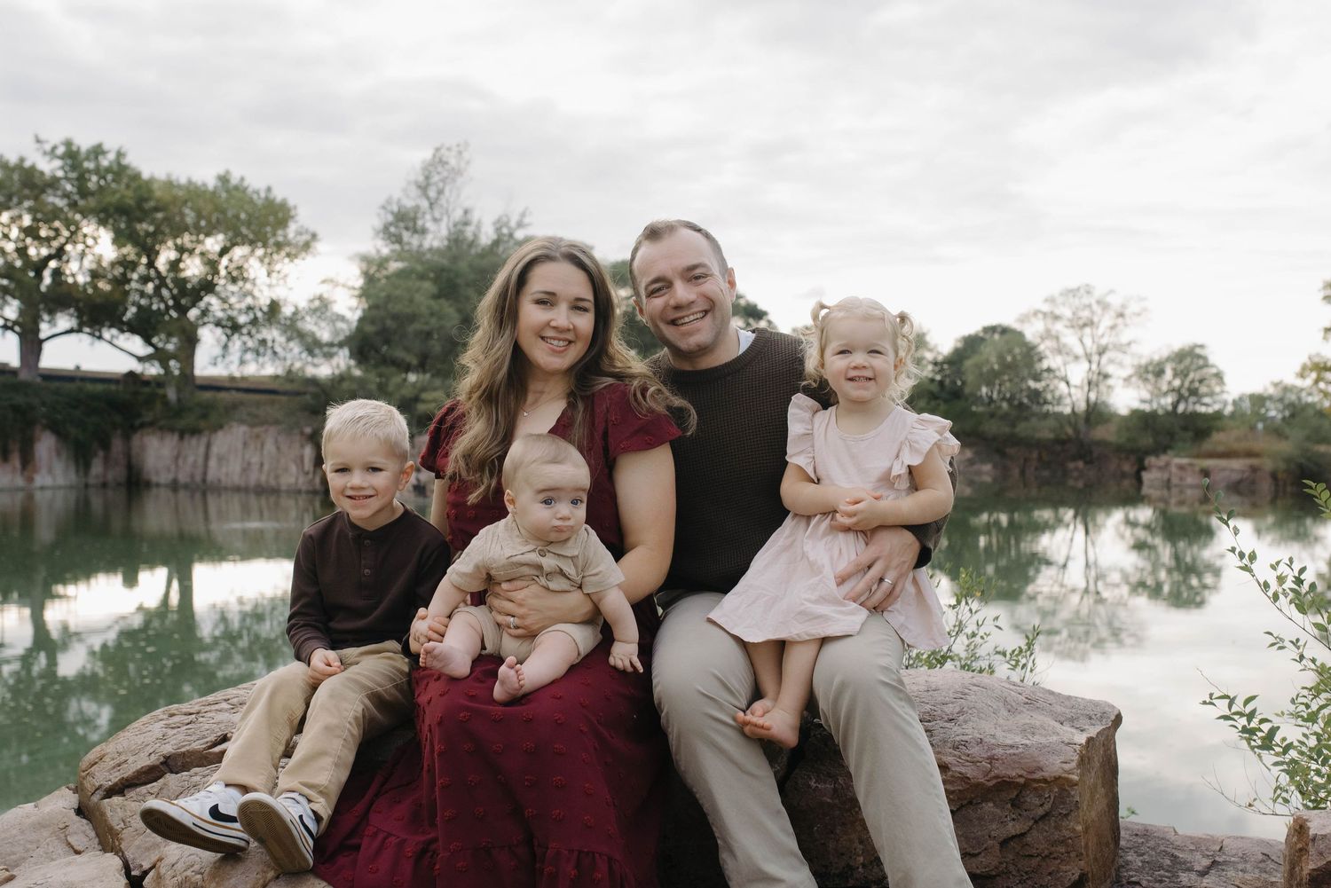 Family of five with three young children sitting together on rocks beside a calm reflective pond.