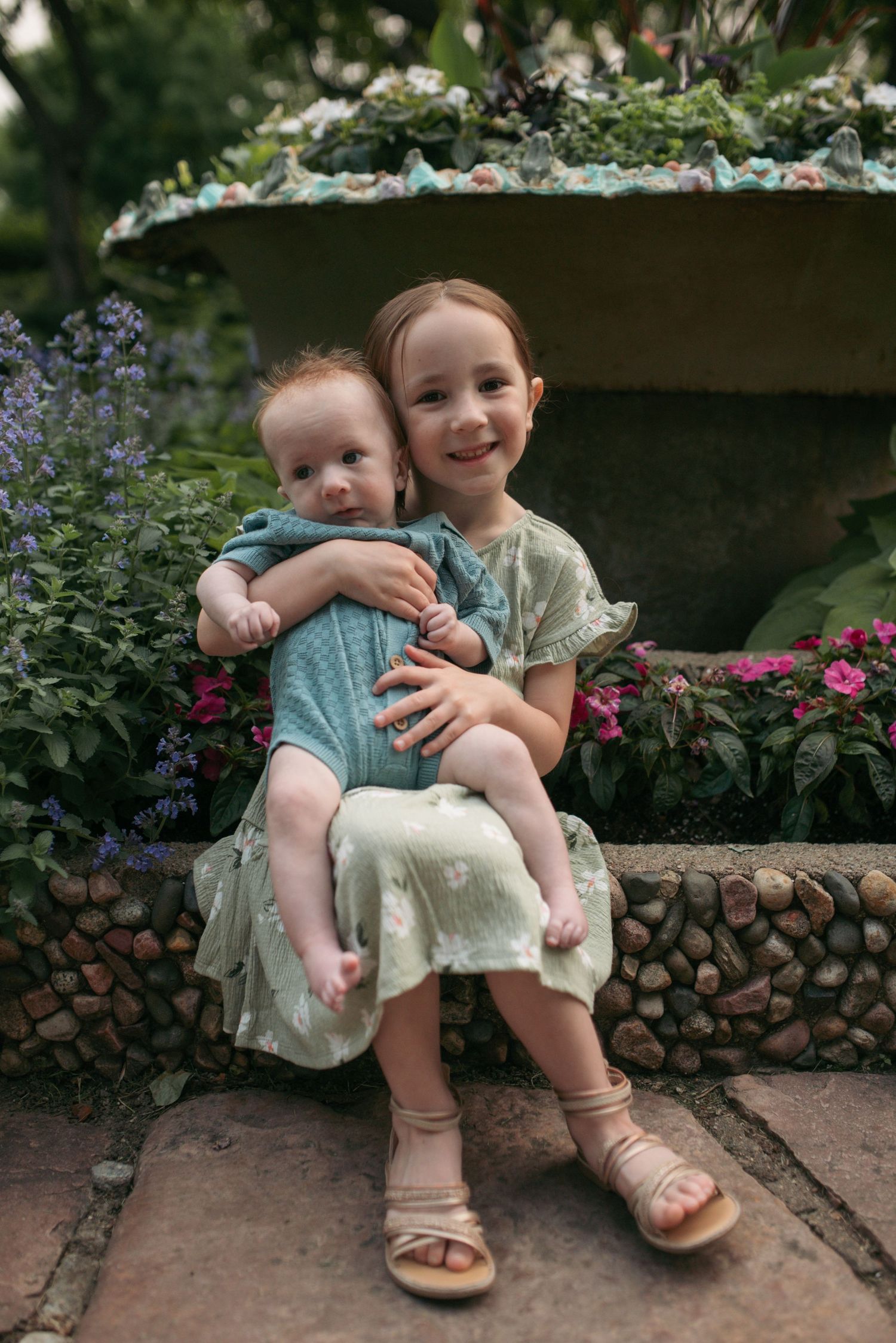 Sweet girl in floral dress hugs baby sibling near colorful garden flowers.